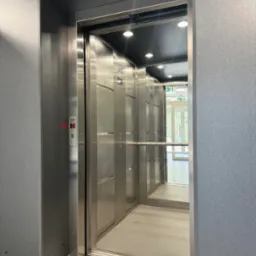 Otwarta, nowoczesna winda ze stali nierdzewnej w budynku z widocznym wyświetlaczem piętra '0' i znakami ewakuacyjnymi.