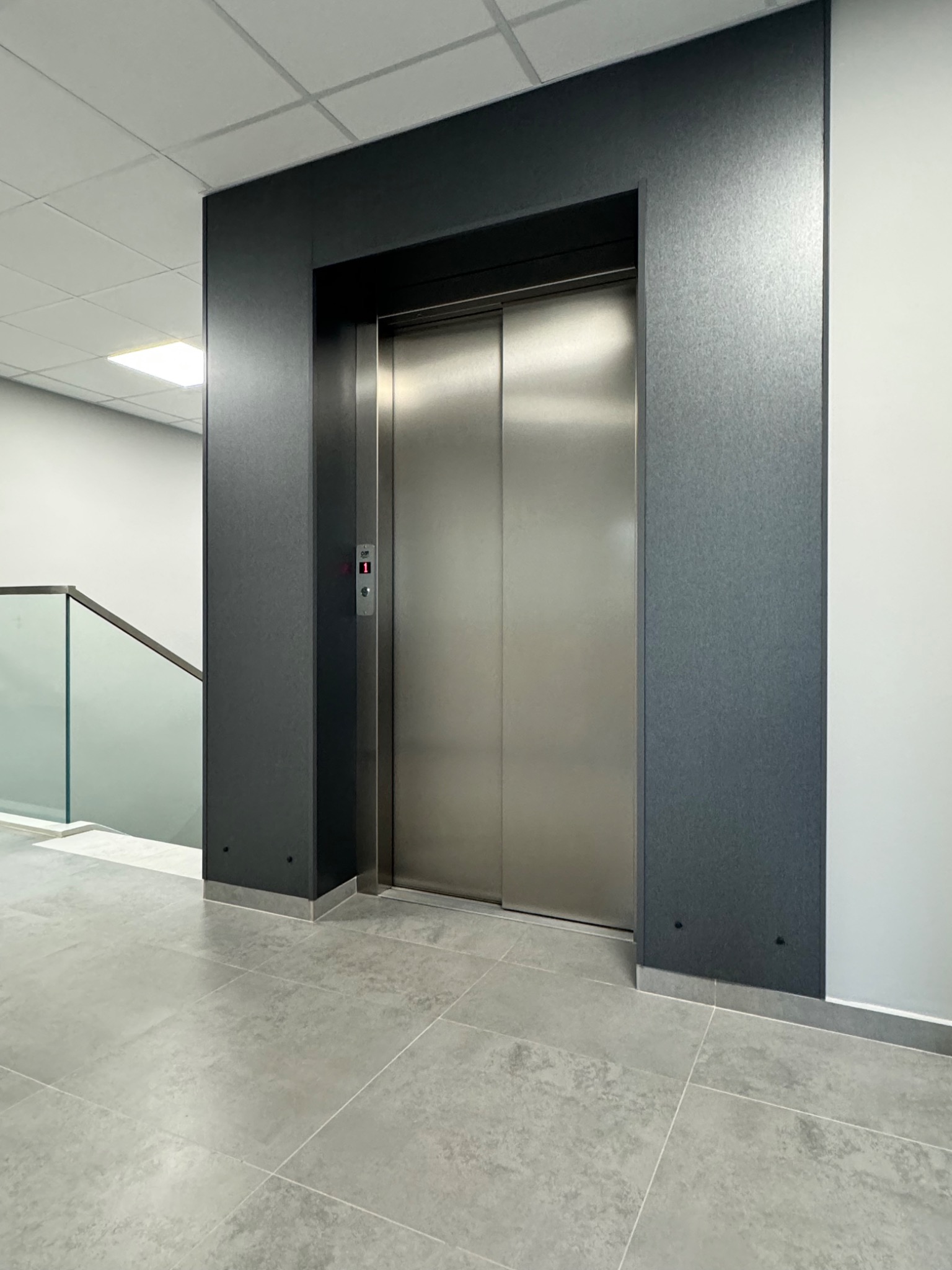 Nowoczesna winda ze stali nierdzewnej z podświetlanym przyciskiem numeru piętra w biurowcu, widok z poziomu podłogi wyłożonej szarymi płytkami.