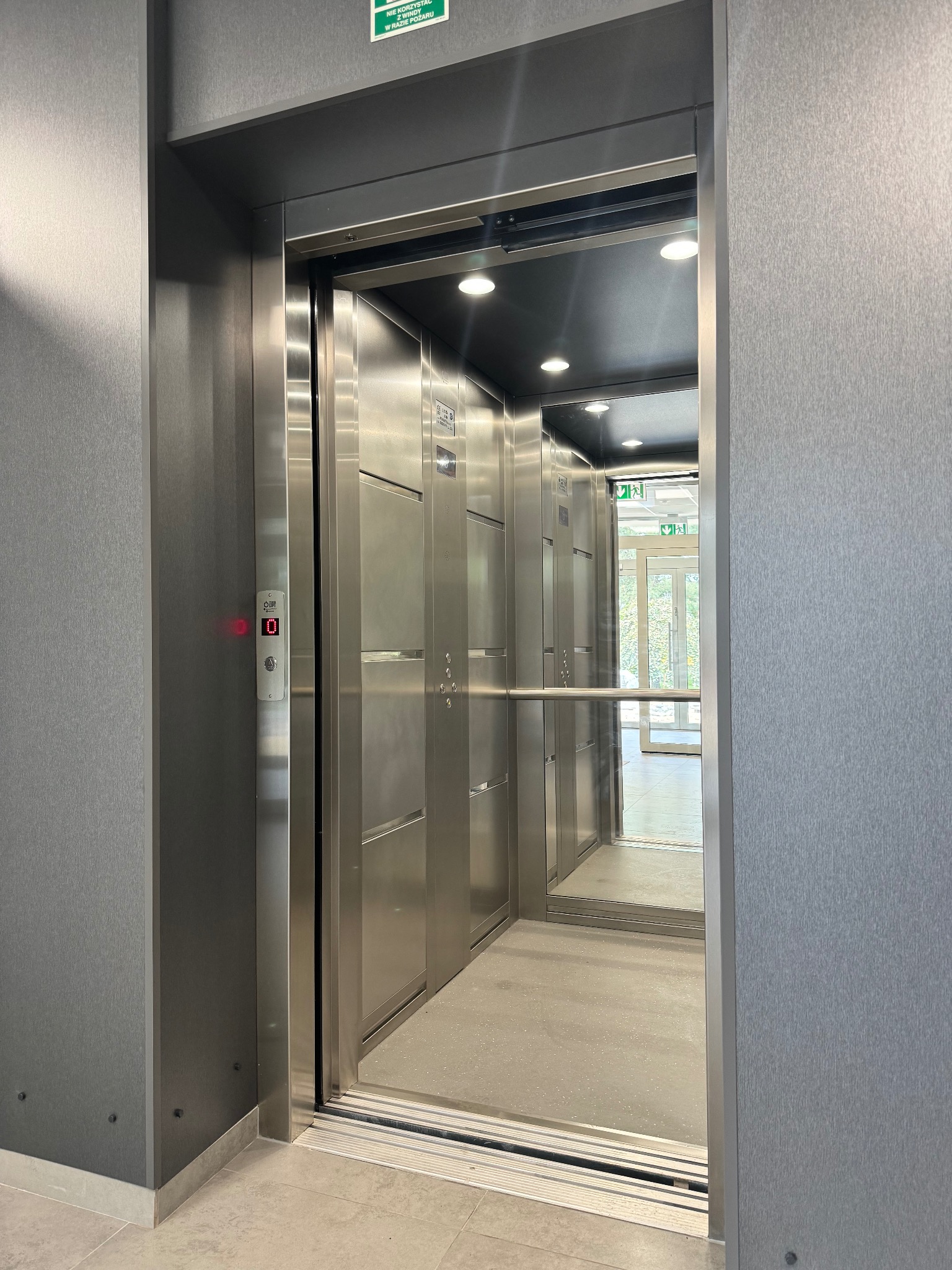 Otwarta, nowoczesna winda ze stali nierdzewnej w budynku z widocznym wyświetlaczem piętra '0' i znakami ewakuacyjnymi.