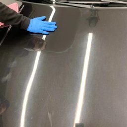 Bomba Garage Auto Detailing - Czarny samochód z widocznymi zarysowaniami na masce, czyszczony przez osobę w niebieskich rękawicach, z użyciem różowej ściereczki.