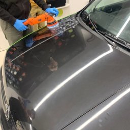Bomba Garage Auto Detailing - Polerowanie czarnego samochodu za pomocą pomarańczowej polerki rotacyjnej; widoczne ślady pasty polerskiej na masce pojazdu, zabezpieczonej zieloną taśmą.