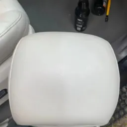 Biały zagłówek samochodowy w trakcie czyszczenia, widoczne ślady użytkowania, czarne rękawice ochronne, butelki z preparatami i pędzel w tle.