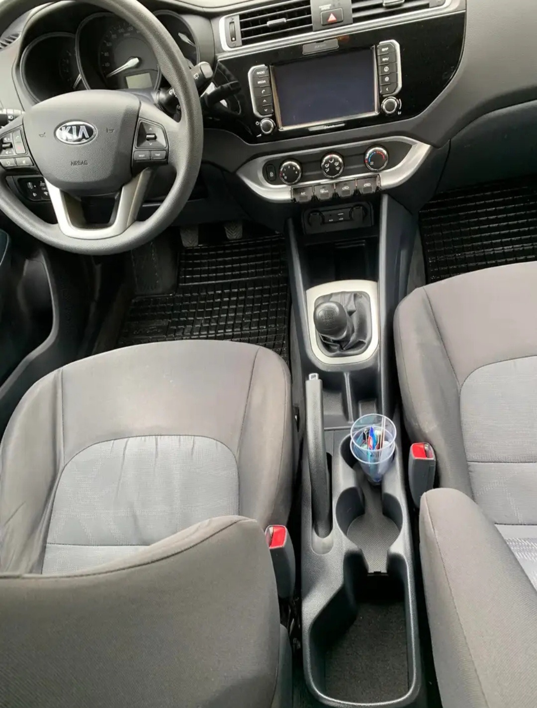 Wnętrze samochodu marki Kia, widok z przodu, obejmujący deskę rozdzielczą, kierownicę, fotele i tunel środkowy z uchwytem na napoje zawierającym długopisy.