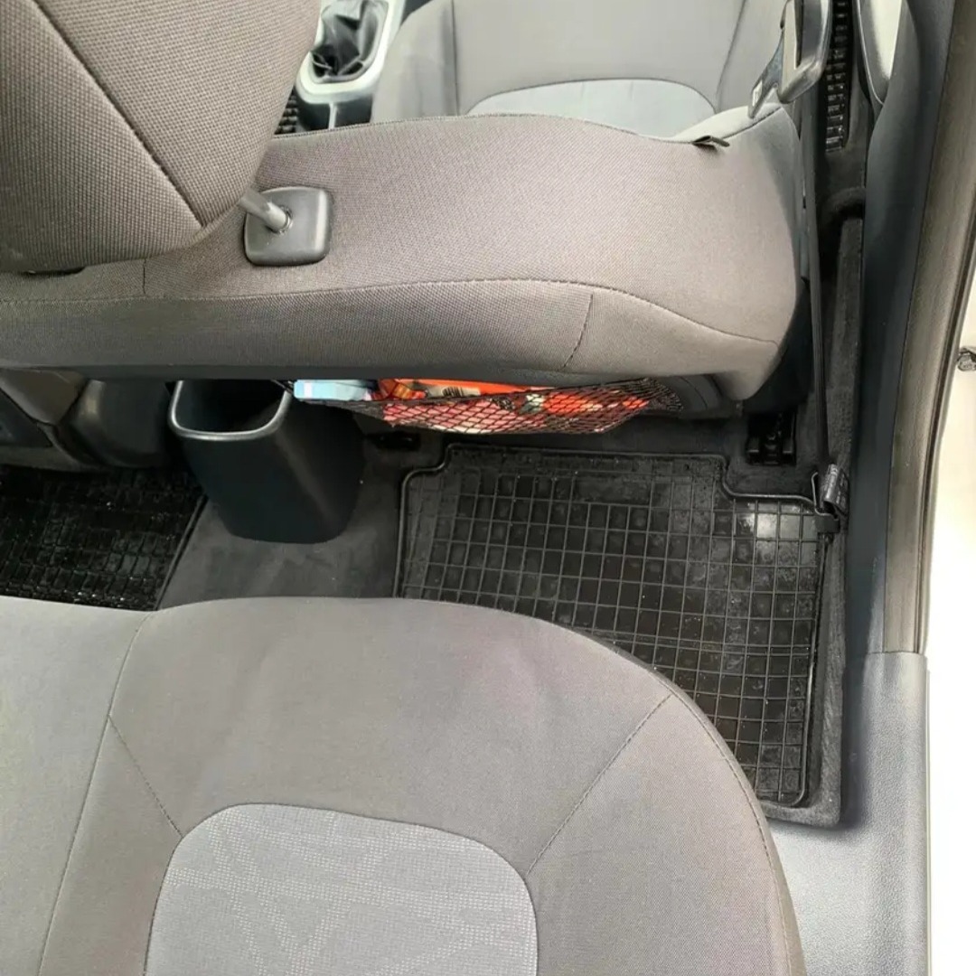 Wnętrze samochodu z widocznymi szarymi fotelami, gumowymi dywanikami z kratką i siatką z pomarańczowymi elementami pod siedzeniem.