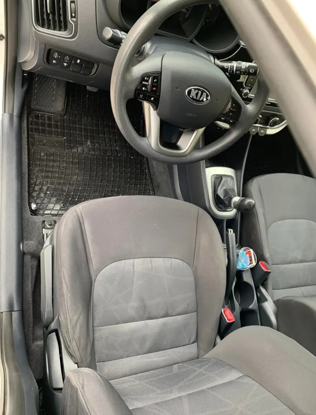Wnętrze samochodu marki Kia, widok z góry na przednie fotele z szarej tkaniny z wzorem, gumowe dywaniki, kierownica z logo Kia, dźwignia zmiany biegów i butelka wody w schowku.