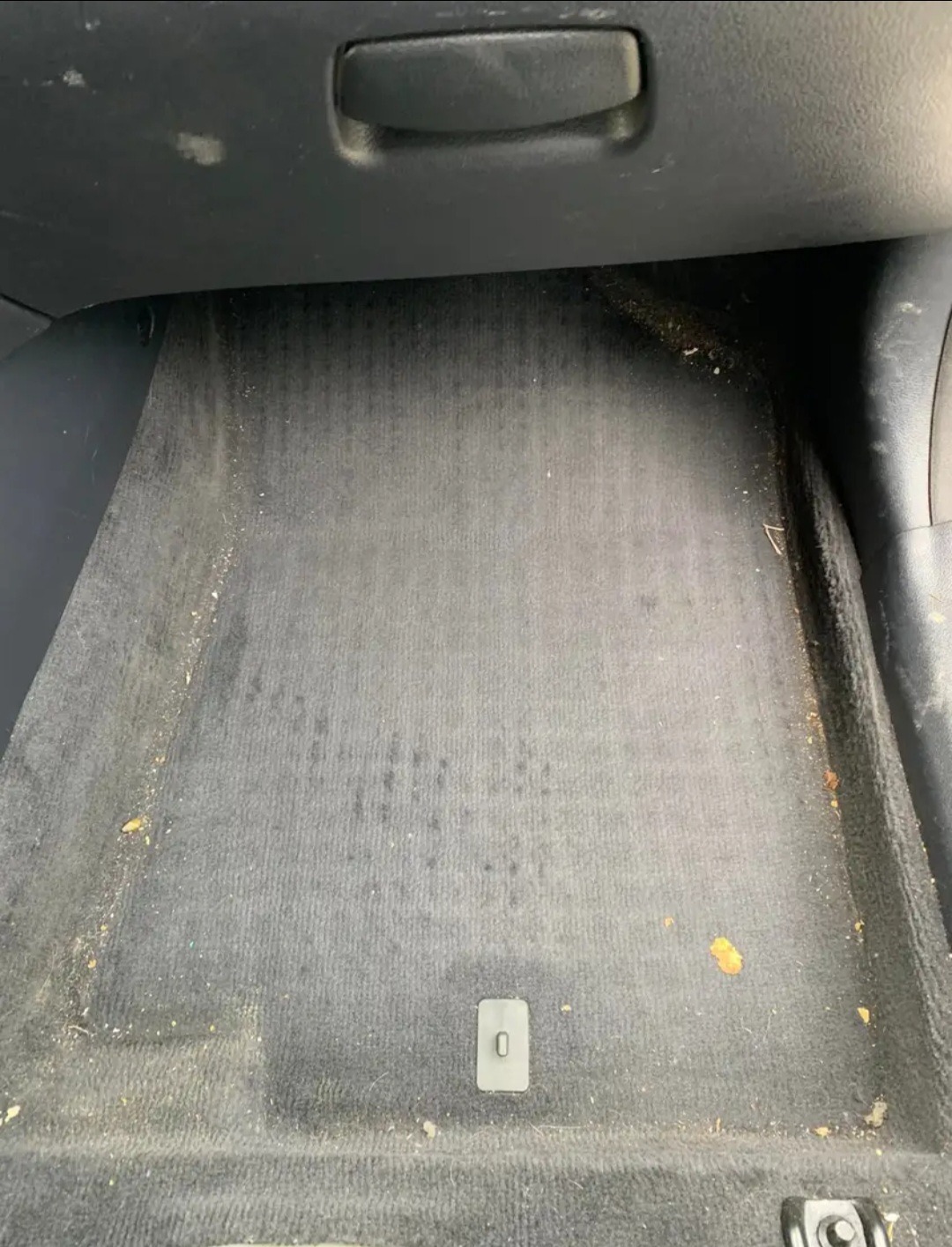 Brudna, szara wykładzina podłogowa w samochodzie z widocznymi plamami, okruchami i drobnymi zabrudzeniami. Widoczny fragment deski rozdzielczej i plastikowych elementów wnętrza.