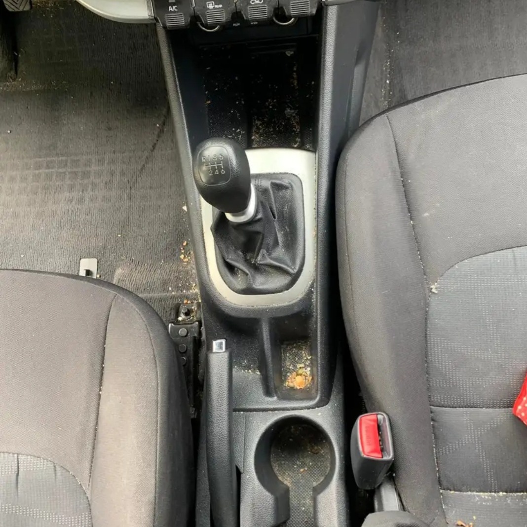 Wnętrze samochodu z widocznymi zabrudzeniami na dywanikach, konsoli środkowej i fotelach, uchwyt na kubek z resztkami, dźwignia zmiany biegów w pozycji neutralnej.