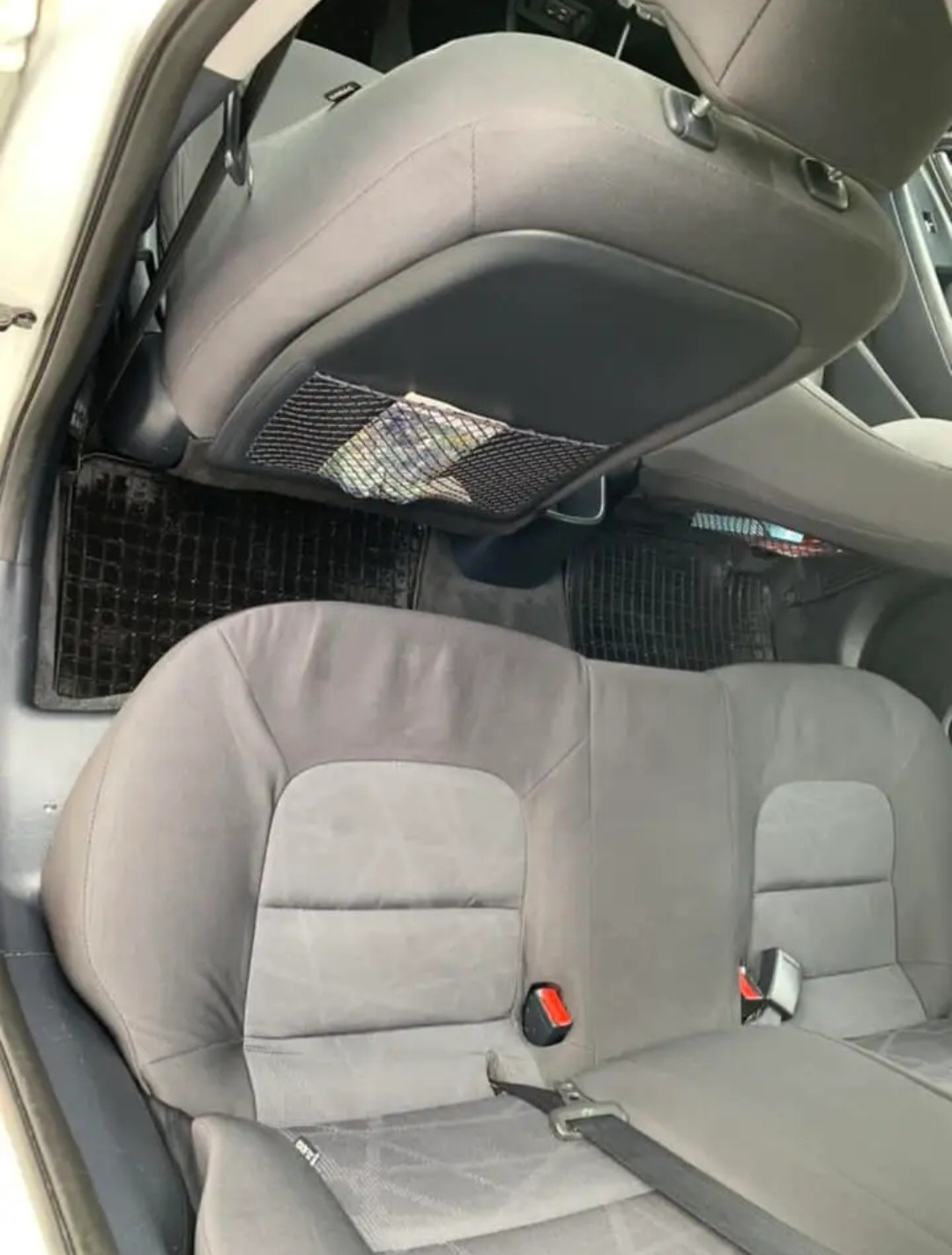 Wnętrze samochodu: tylna kanapa z szarej tapicerki, czarne dywaniki pod nogami pasażerów, siatka na oparciu przedniego fotela z widoczną zawartością.