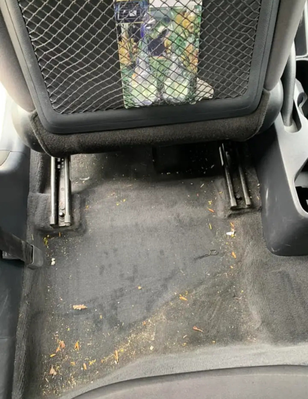Brudna podłoga samochodu przed czyszczeniem, widoczne resztki organiczne, piasek i kurz, z metalowymi szynami fotela i siatką na oparciu fotela.