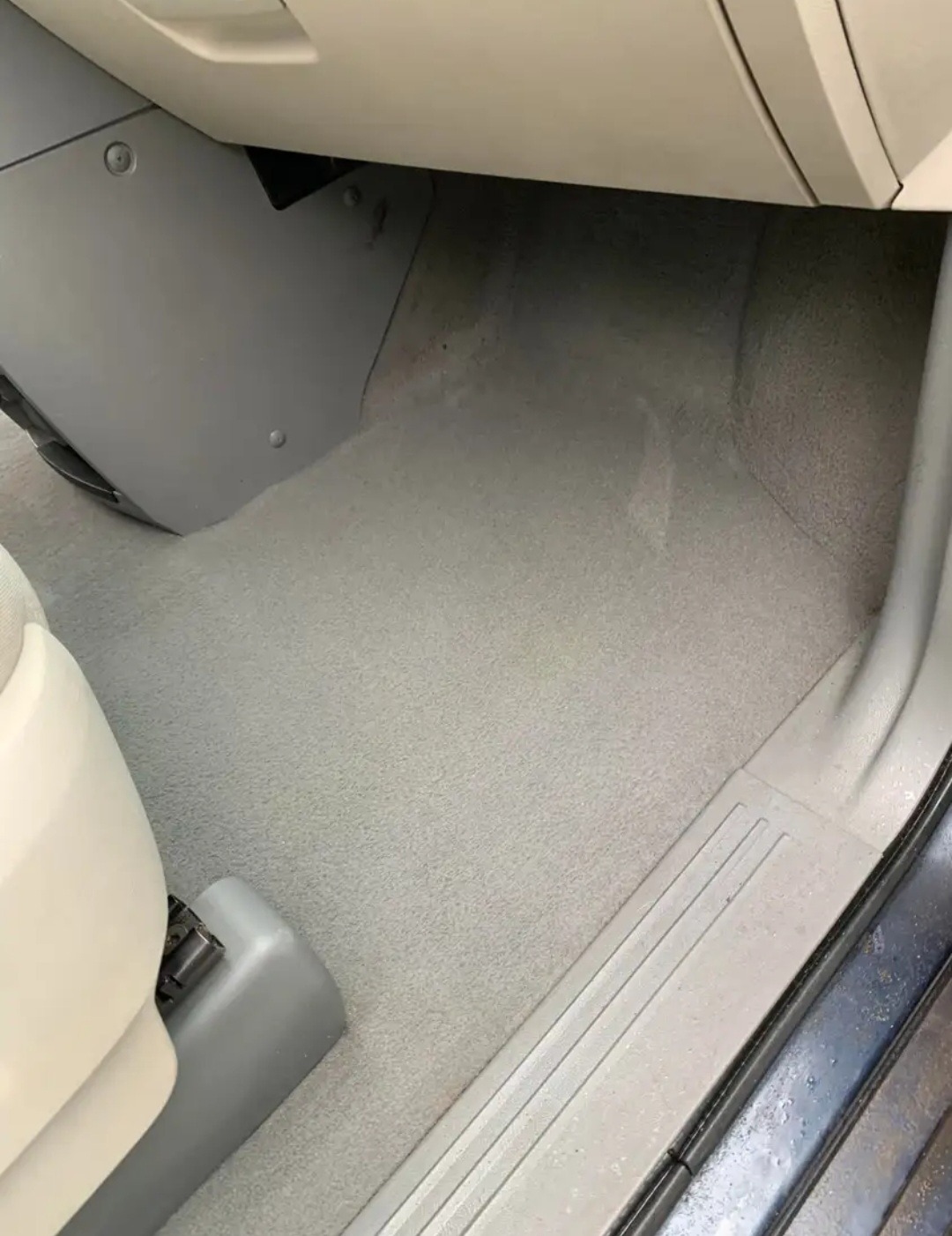 Wnętrze samochodu, widok na dywanik pod nogami kierowcy z fragmentem fotela, listwy progowej i dolnej części deski rozdzielczej. Dywanik w kolorze beżowym, lekko przybrudzony.