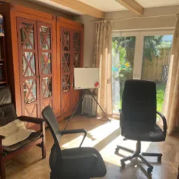 Gabinet z drewnianymi meblami, tablicą suchościeralną i dwoma krzesłami, widok na ogród przez okno balkonowe z zasłonami.