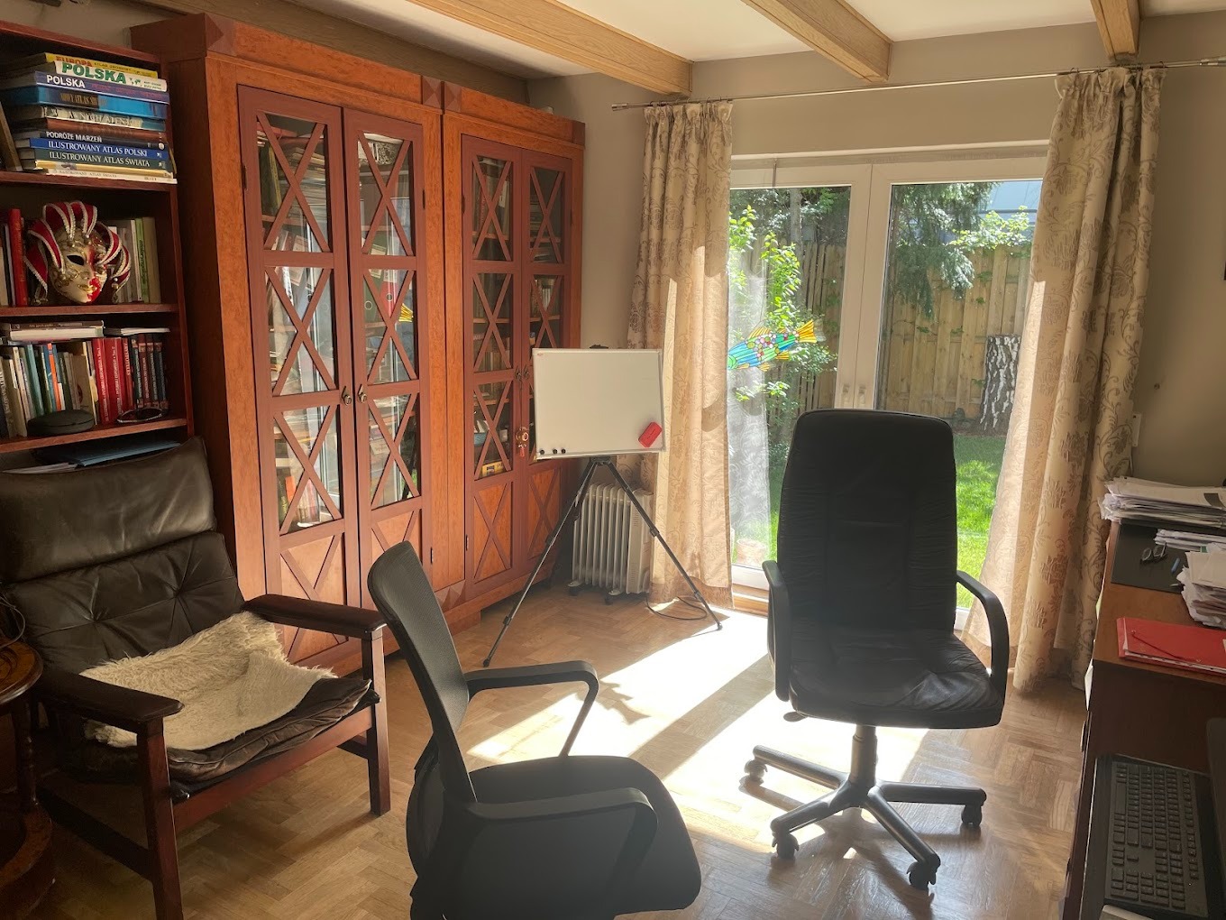 Gabinet z drewnianymi meblami, tablicą suchościeralną i dwoma krzesłami, widok na ogród przez okno balkonowe z zasłonami.
