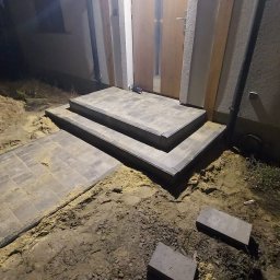 Figur-Bruk - Schody wejściowe z kostki brukowej prowadzące do drzwi, widoczne świeżo ułożone płyty chodnikowe i nasypana ziemia po bokach, wykonane w nocy.