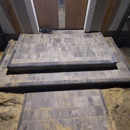 Figur-Bruk - Schody wejściowe do domu wyłożone szarą kostką brukową, widoczne ślady piasku na powierzchni i wokół, kabel przed drzwiami.