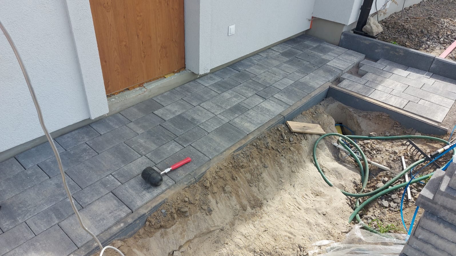 Ułożona szara kostka brukowa przed wejściem do domu, widoczny gumowy młotek i wykop z przewodami, etap prac wykończeniowych.