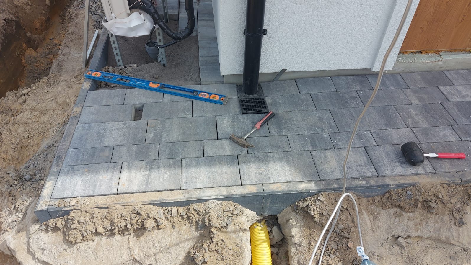 Fragment posesji w trakcie prac brukarskich: ułożona część kostki brukowej, poziomica, młotek, gumowy młotek, żółta rura w wykopie, widoczny grunt.