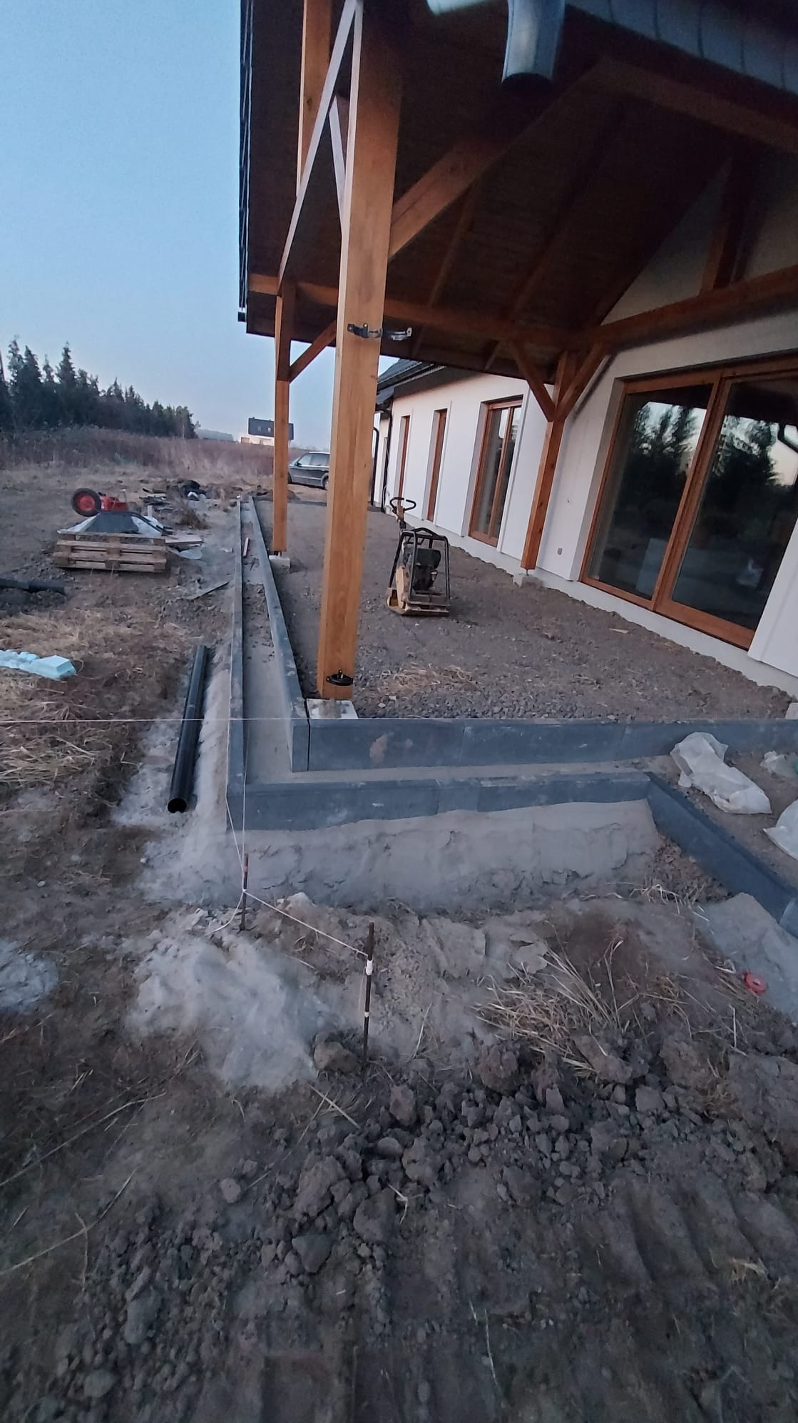 Przygotowanie podłoża pod taras: widoczne obrzeża z szarej kostki, zagęszczarka wibracyjna oraz paleta z walcem drogowym w tle, teren w trakcie prac ziemnych.