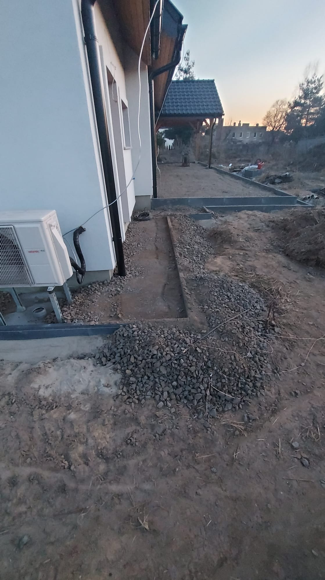 Przygotowany teren pod chodnik z obrzeżami, częściowo wysypany grysem, widoczny budynek z klimatyzatorem na ścianie i zadaszenie w tle, etap prac ziemnych.