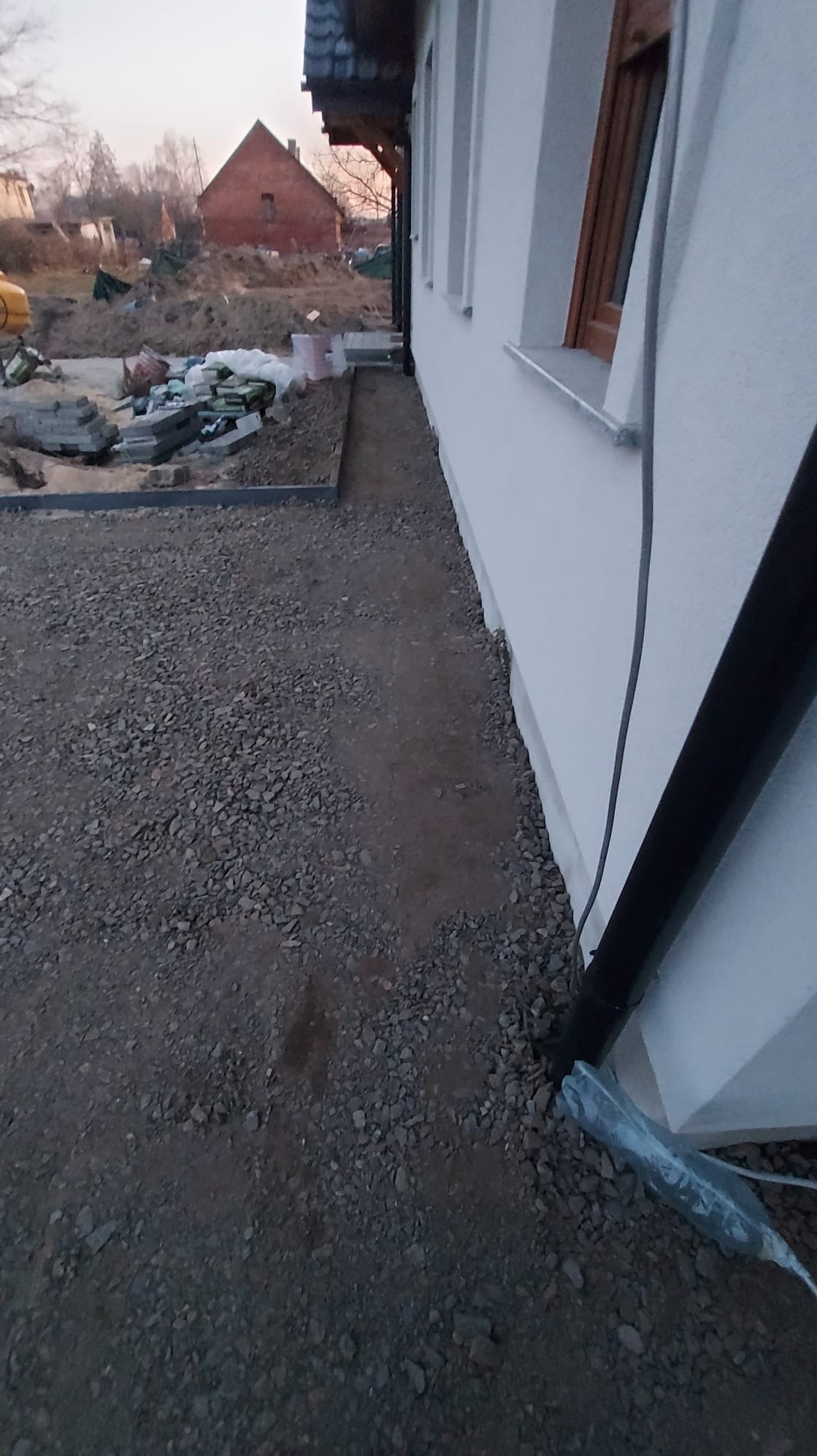 Przygotowanie terenu pod kostkę brukową: widok na warstwę podsypki żwirowej przy ścianie budynku z zamontowaną rynną spustową, w tle widoczny stos kostki i koparka.