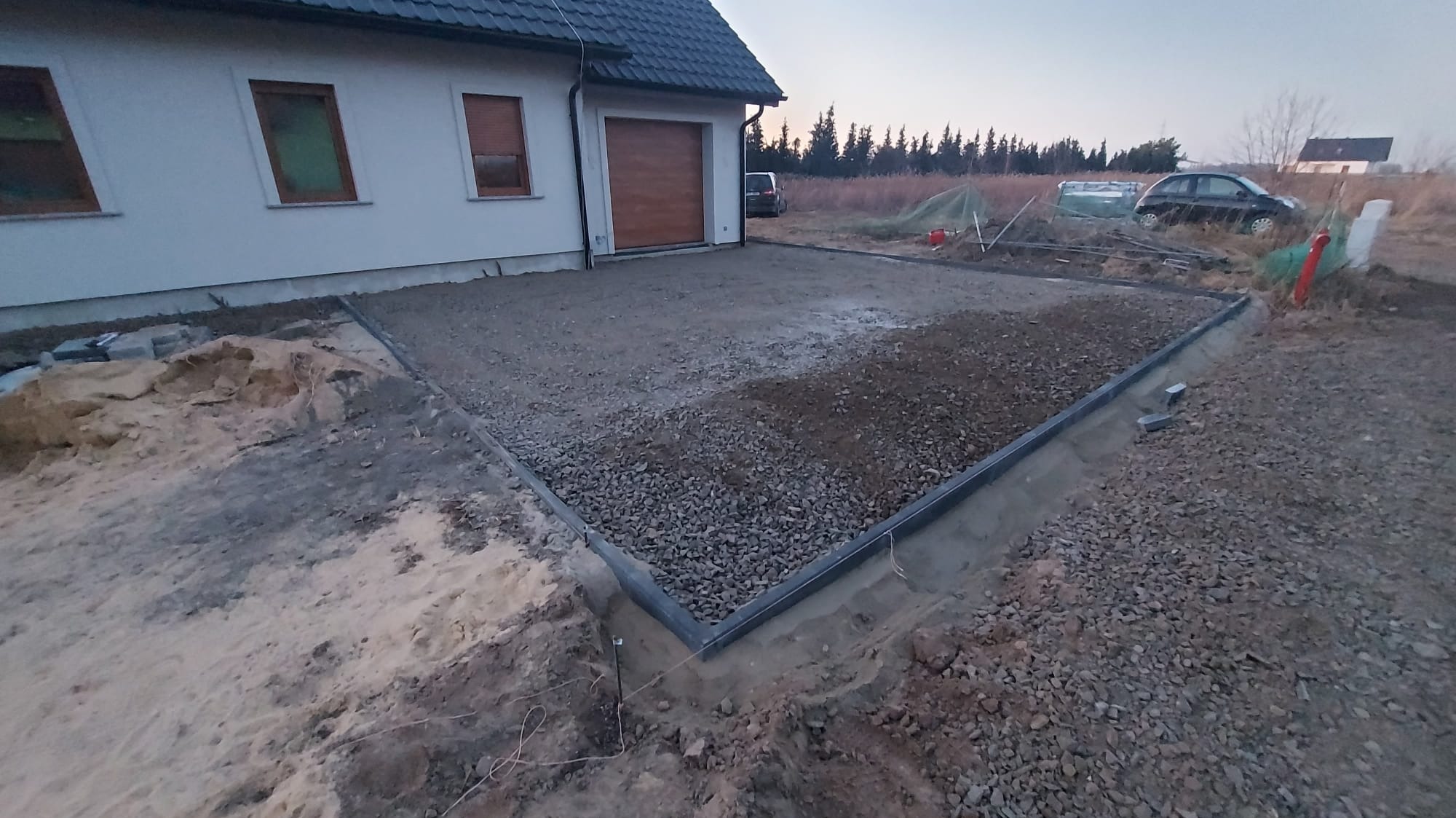 Przygotowany teren pod kostkę brukową: wyrównany grunt z obrzeżami, częściowo wysypany żwir, widok na dom z garażem i samochody w tle, szarobłękitne, pochmurne niebo.