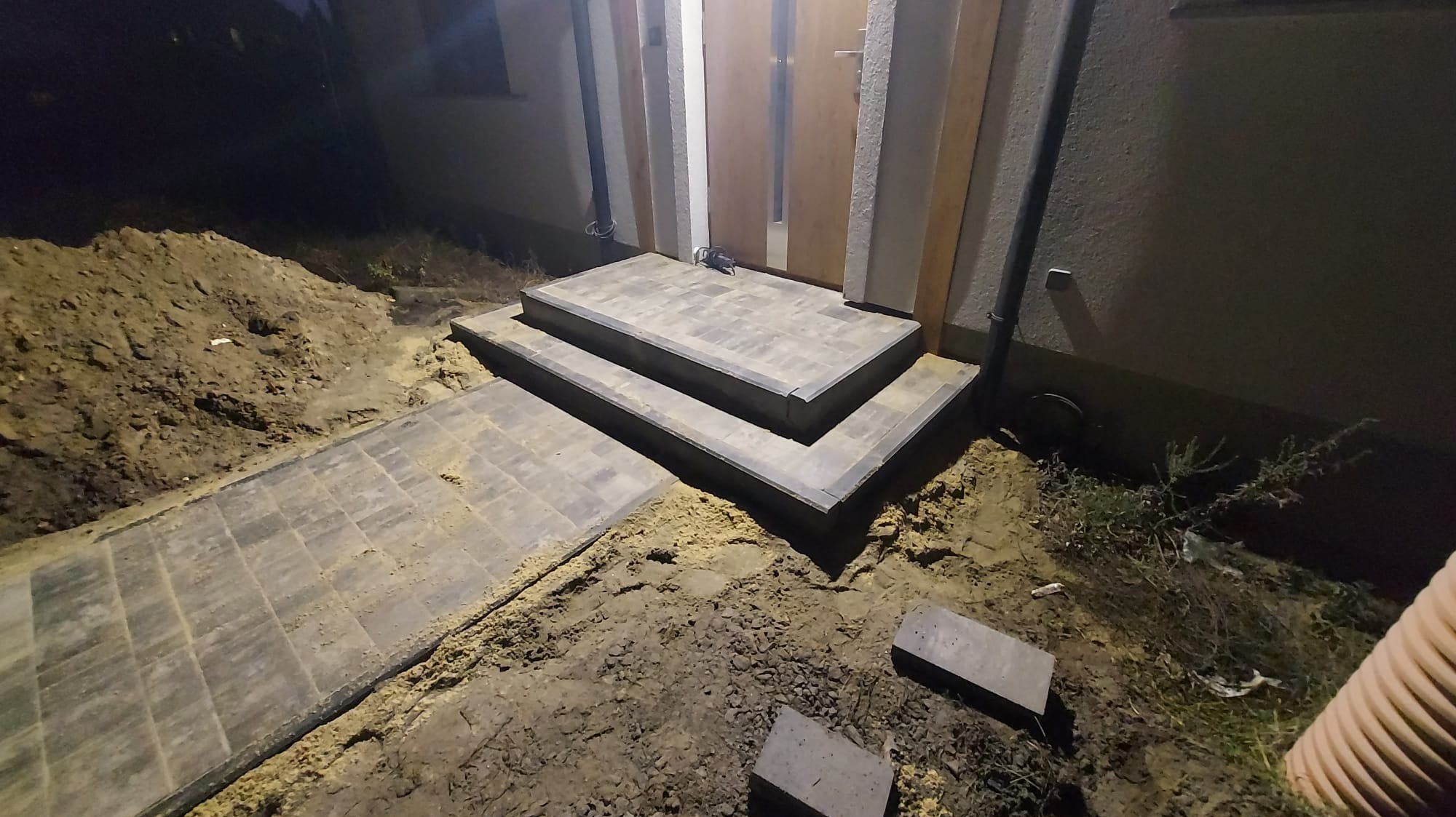 Schody wejściowe z kostki brukowej prowadzące do drzwi, widoczne świeżo ułożone płyty chodnikowe i nasypana ziemia po bokach, wykonane w nocy.
