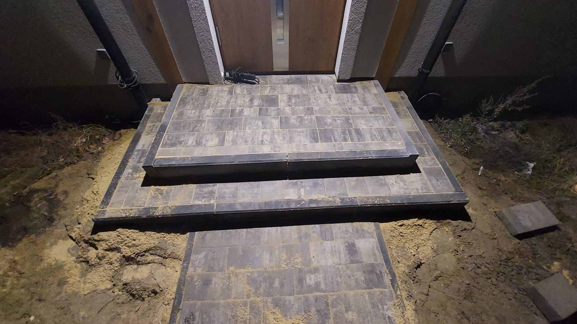 Schody wejściowe do domu wyłożone szarą kostką brukową, widoczne ślady piasku na powierzchni i wokół, kabel przed drzwiami.