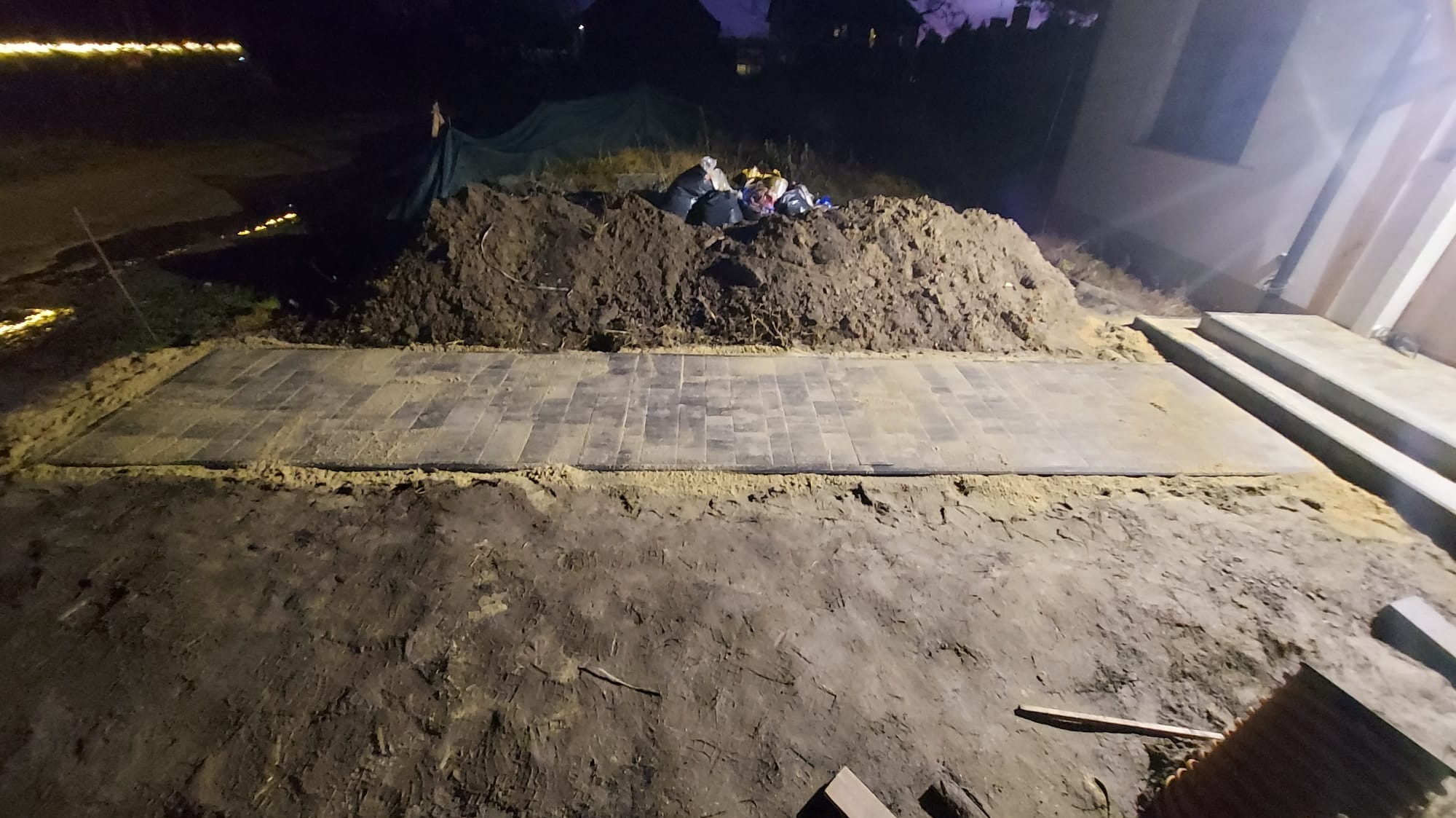 Fragment ułożonej, prostokątnej powierzchni z szarej kostki brukowej, obrzeża z piasku, obok pryzma ziemi z workami na śmieci, nocne oświetlenie.