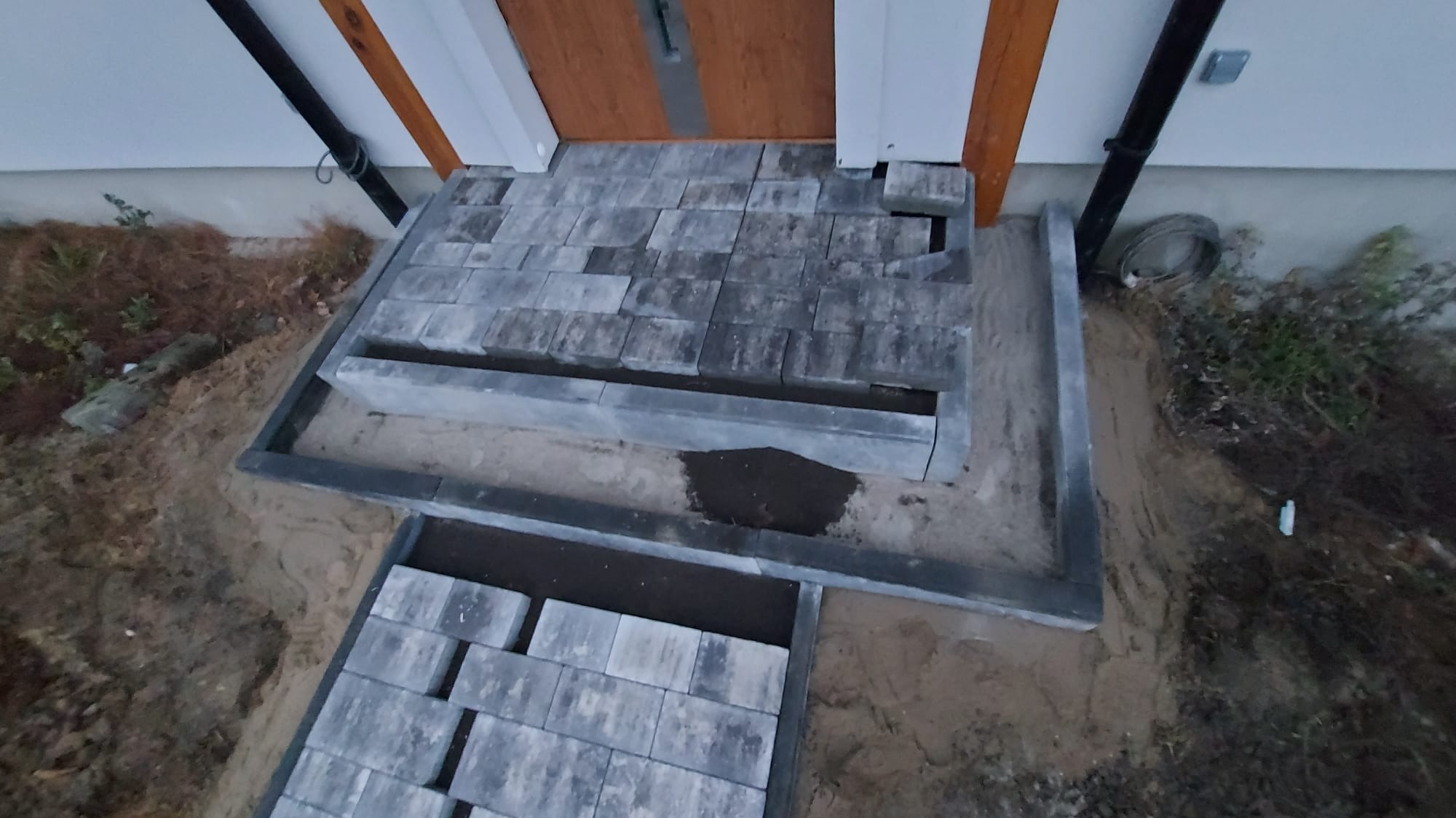 Fragment układania kostki brukowej przed wejściem do domu, widoczne niedokończone ułożenie kostki i obrzeża w piasku.