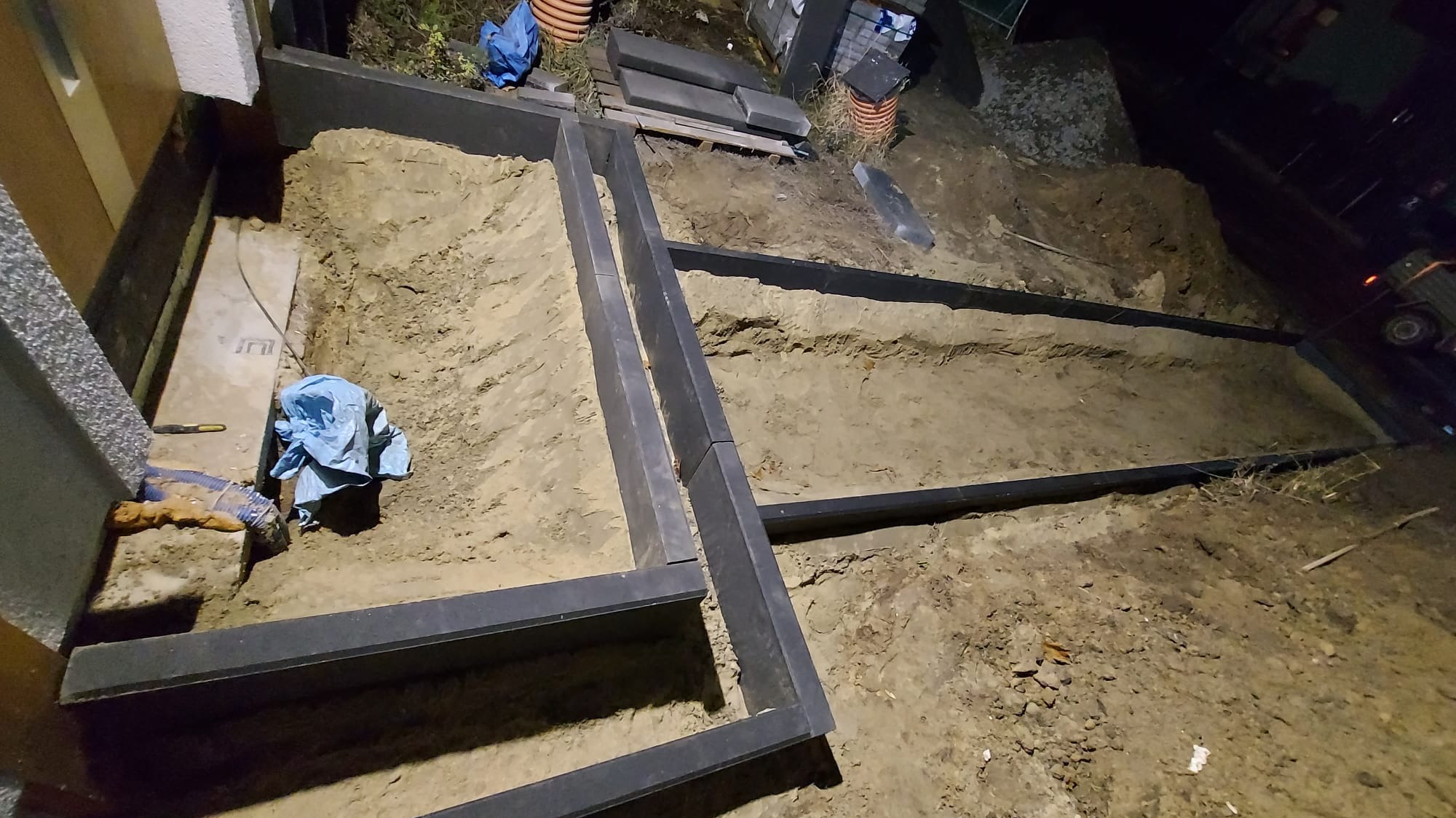 Przygotowany teren pod przyszłą nawierzchnię, widoczne obramowanie z elementów betonowych, piasek, rury drenarskie i częściowo ułożone płyty.