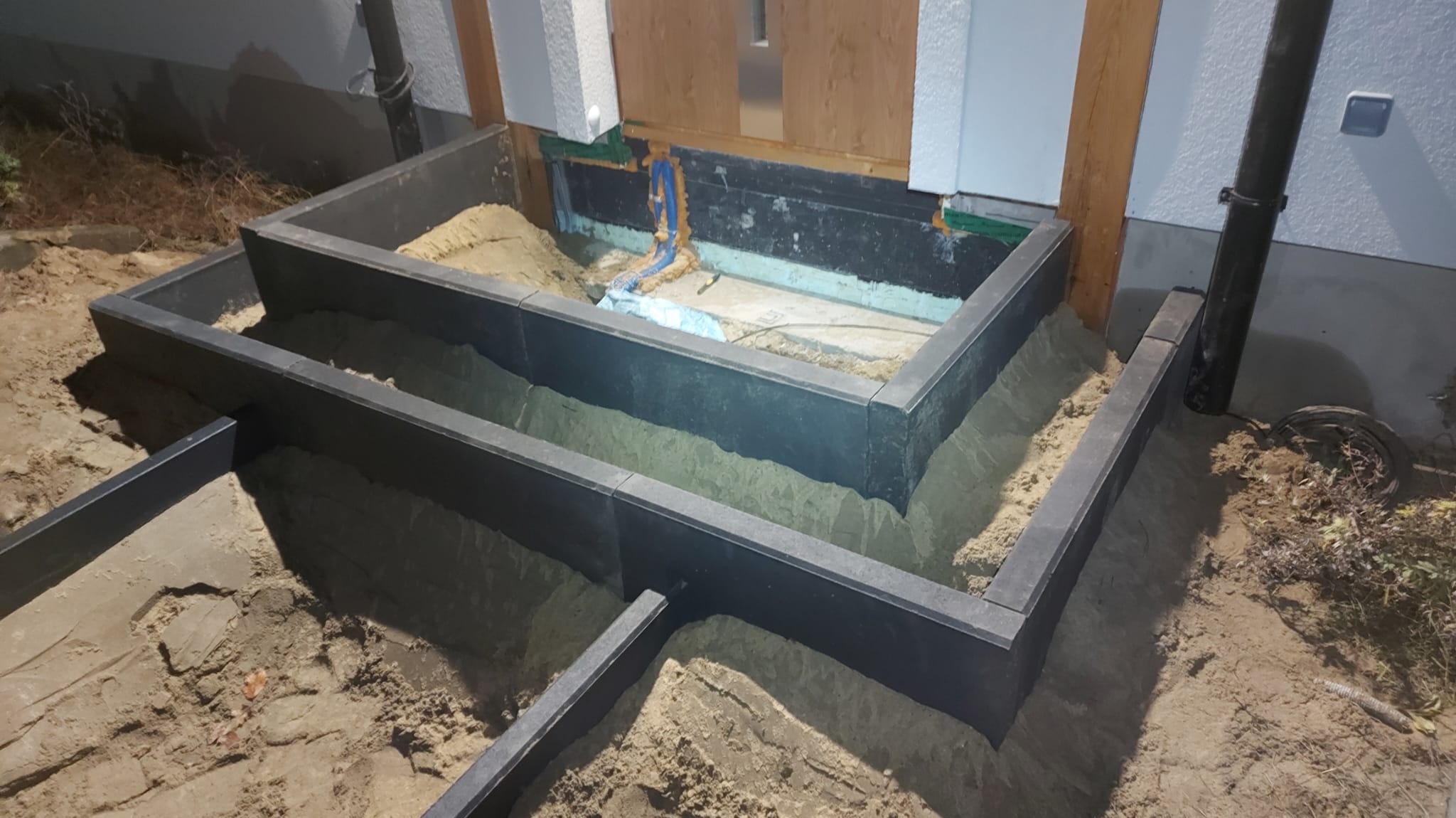 Przygotowanie podłoża pod kostkę brukową: czarne obrzeża ułożone w kilku poziomach, wypełnione piaskiem, widoczne instalacje przy ścianie budynku.