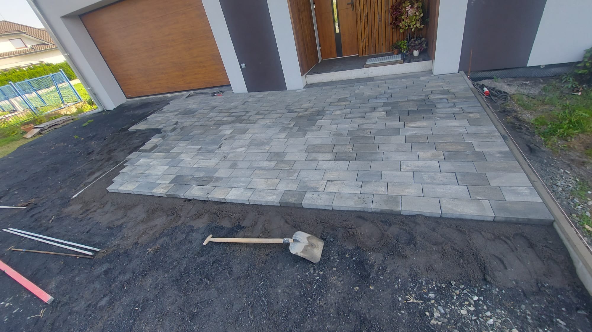 Częściowo ułożona szara kostka brukowa przed nowoczesnym domem z brązową bramą garażową i drewnianymi drzwiami wejściowymi, widoczna łopata i poziomice na czarnej ziemi.