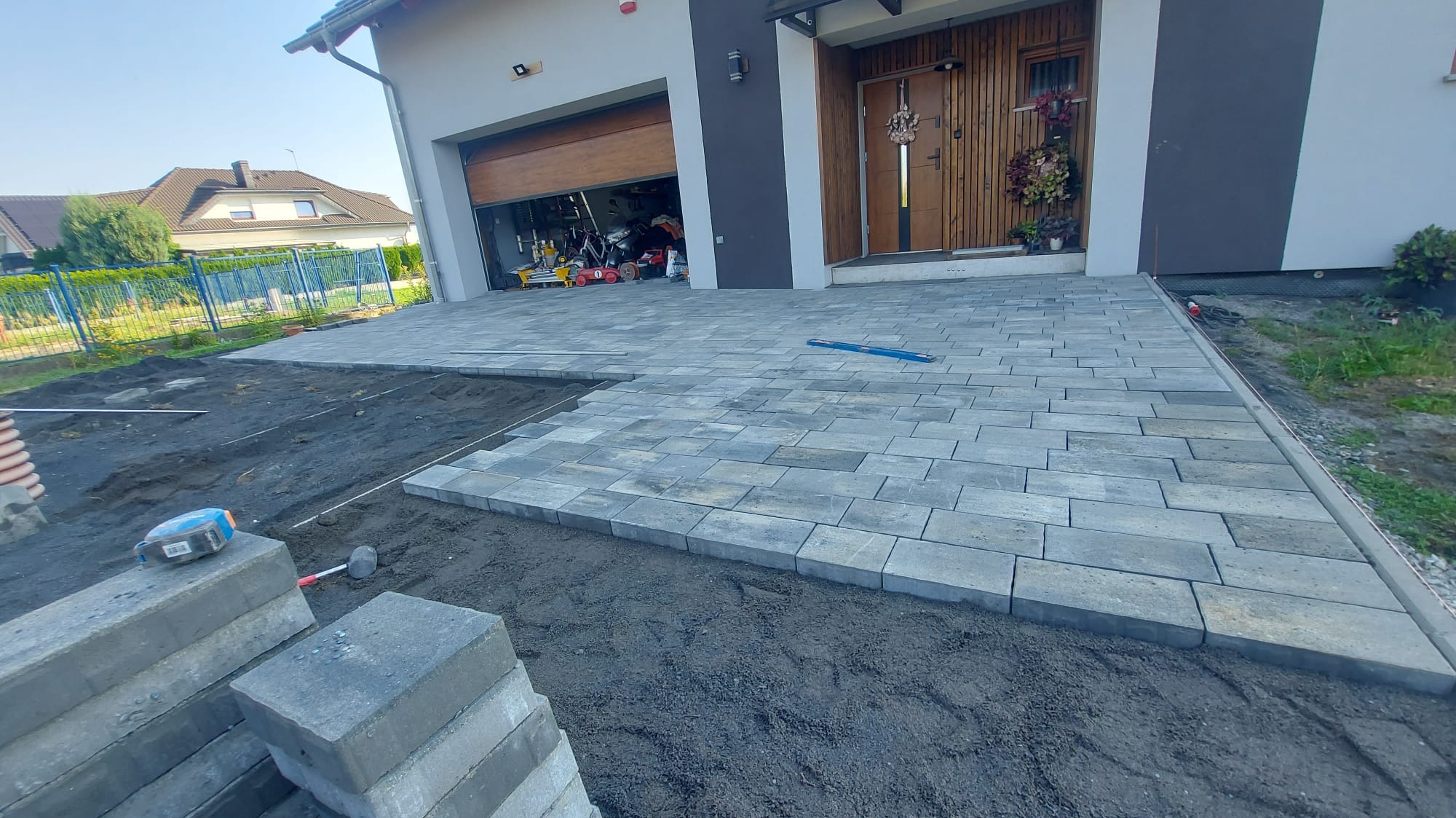 Fragment podjazdu z szarej kostki brukowej w trakcie układania, widoczny piasek podsypkowy, w tle dom z otwartym garażem i narzędziami.
