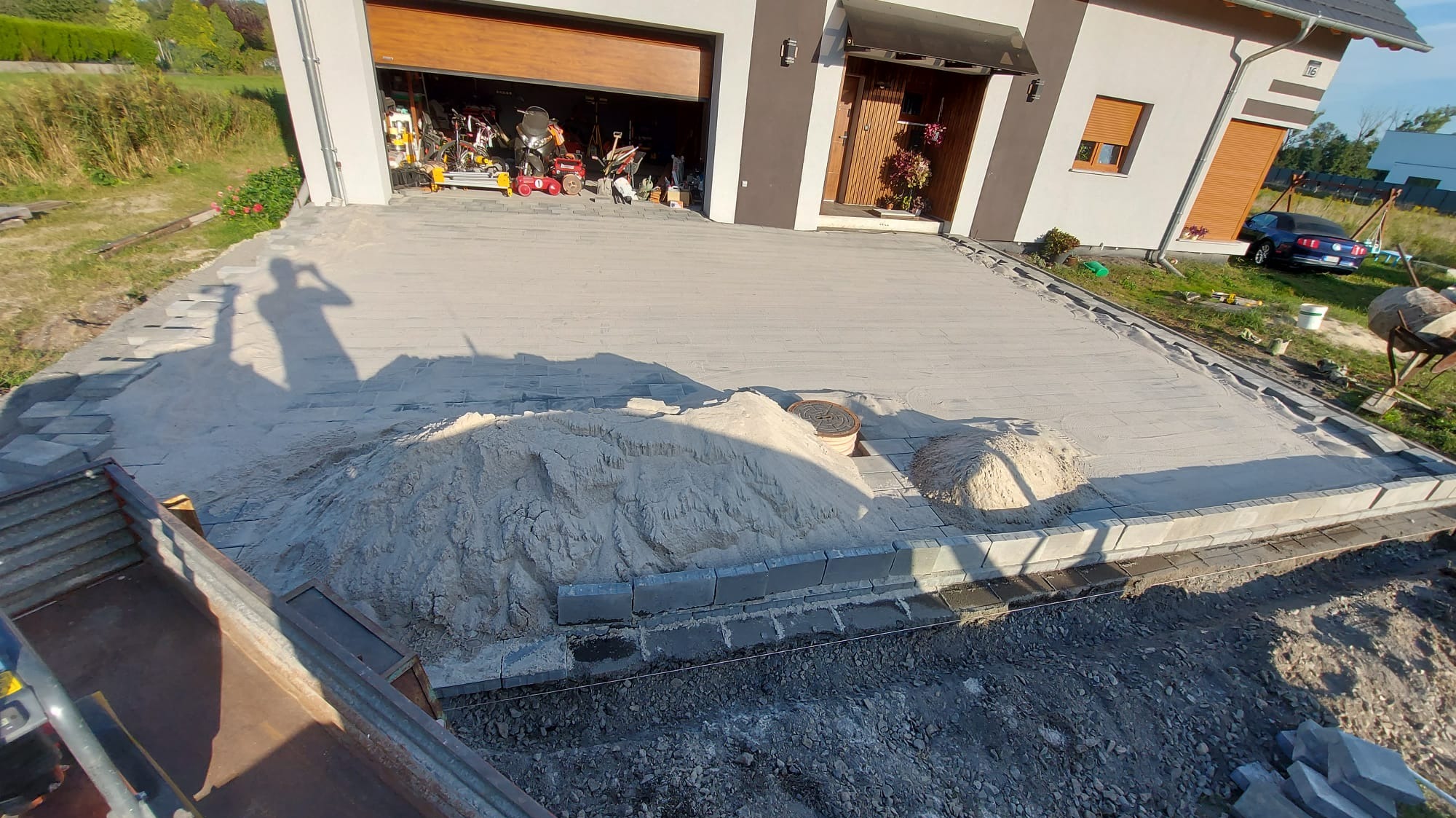 Przygotowanie podłoża pod kostkę brukową na podjeździe do domu jednorodzinnego; widoczny piasek, obrzeża z kostki, cień osoby robiącej zdjęcie oraz otwarty garaż z narzędziami w tle.