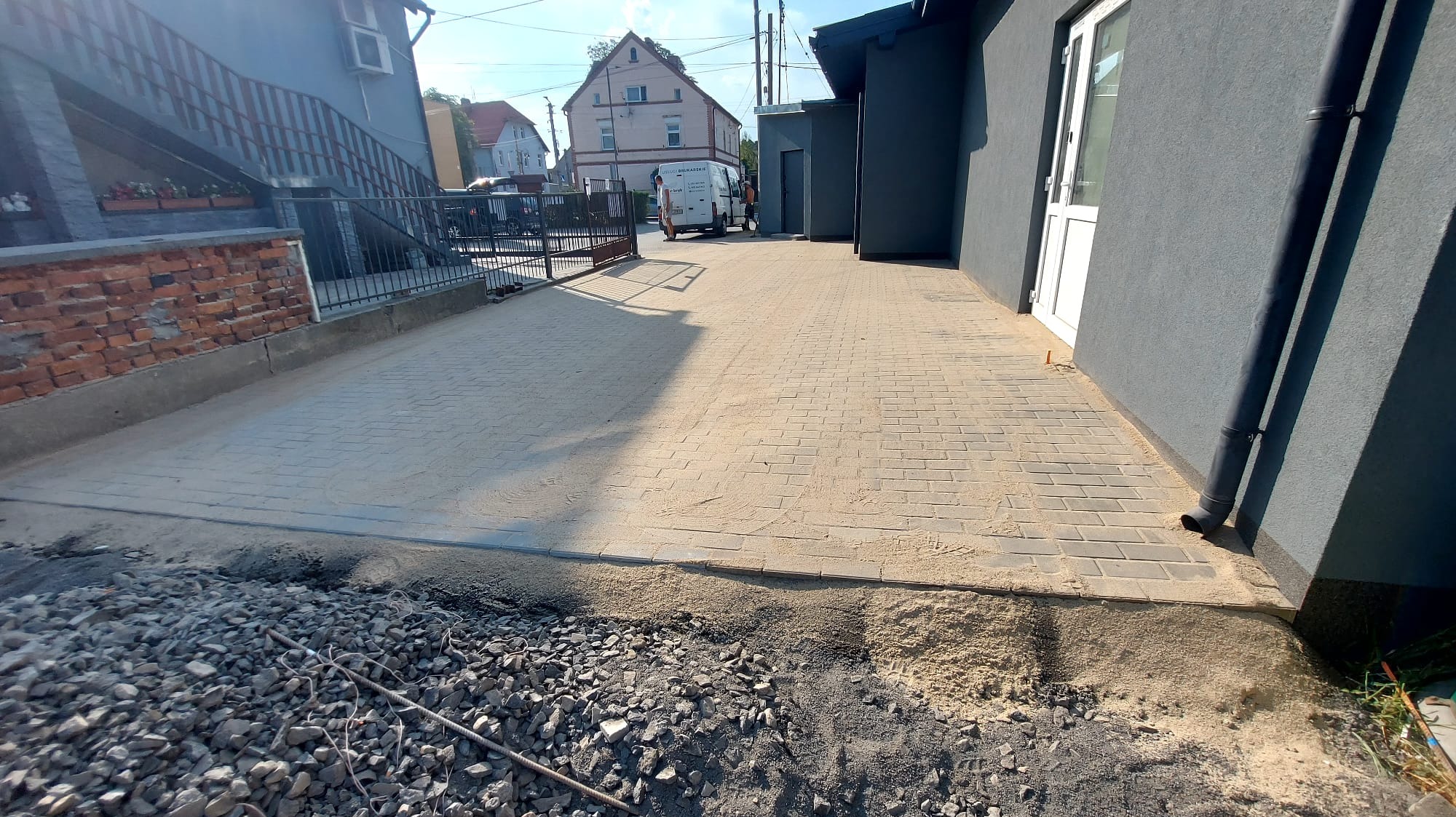 Nowo ułożona, jasna kostka brukowa na podjeździe, częściowo zasypana piaskiem, z widocznym stosem kamieni i żwiru w dolnej części kadru, sugerującym trwające prace wykończeniowe.