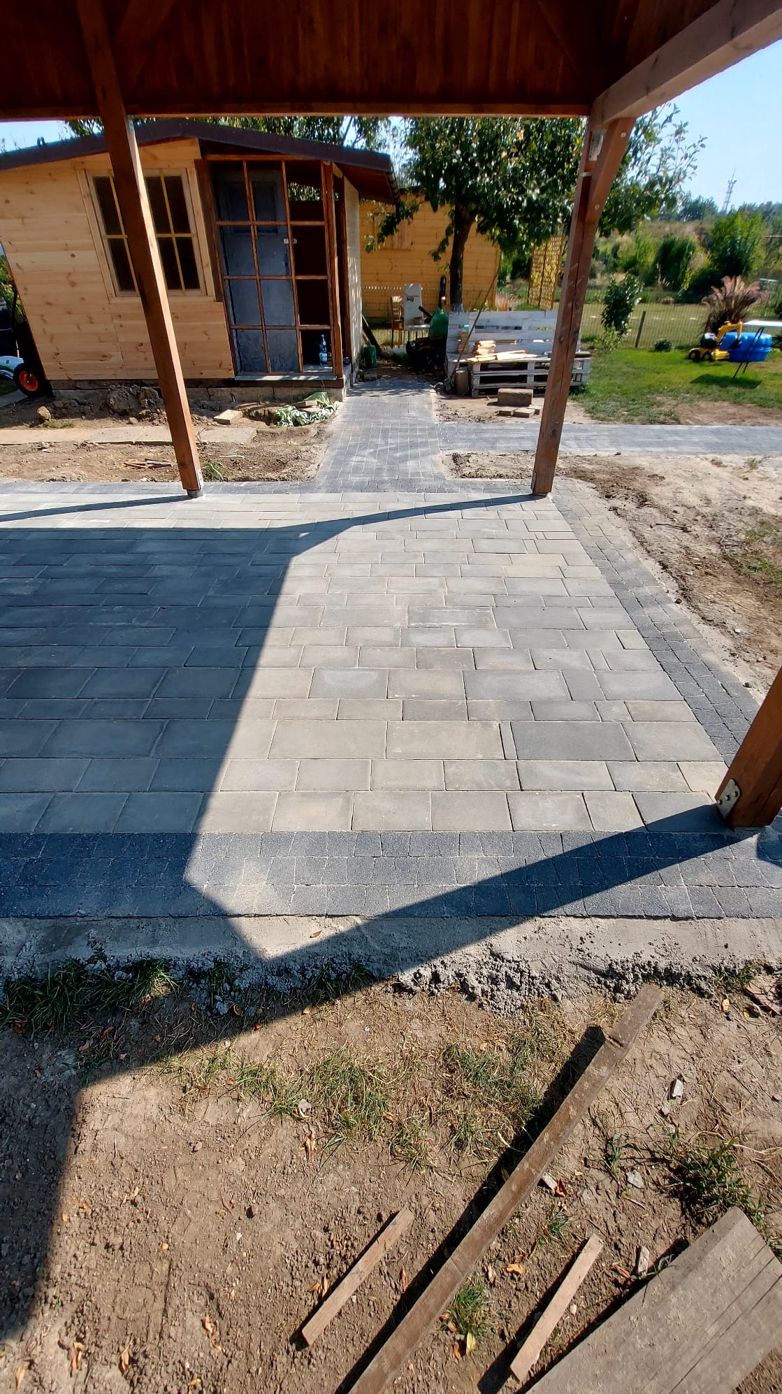 Ułożona szara kostka brukowa na posesji z drewnianą altaną i domkiem narzędziowym w tle, widoczne nierówności terenu i materiały budowlane.
