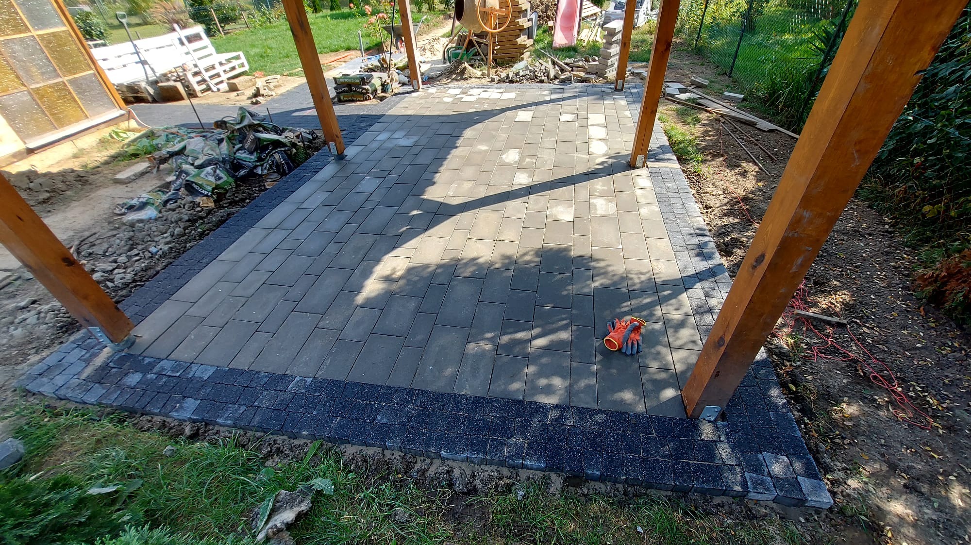 Nowo ułożona, szara kostka brukowa na tarasie z drewnianą konstrukcją z czterema słupami, obrzeże z ciemniejszej kostki, pomarańczowe rękawice robocze leżą na powierzchni.