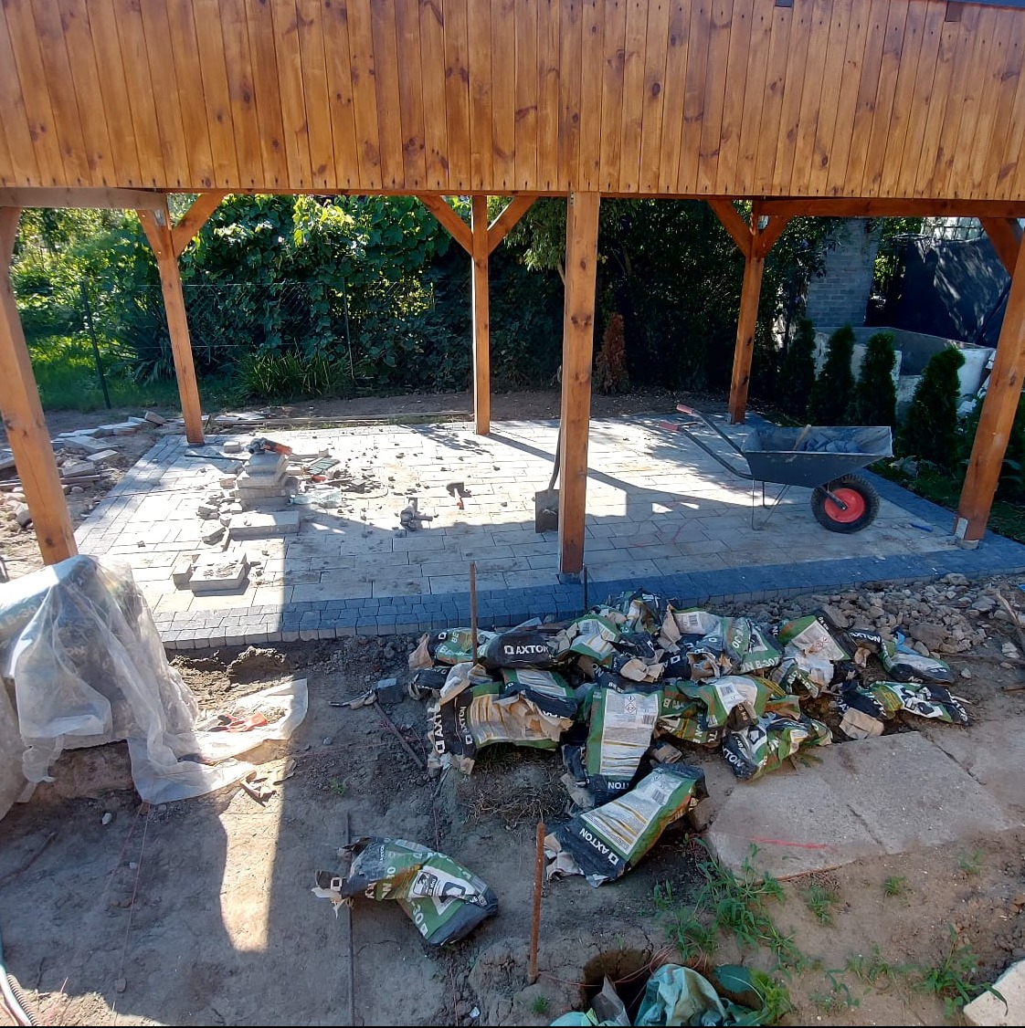 Budowa zadaszenia z kostki brukowej w ogrodzie, widoczne porozrzucane worki z zaprawą Axton, taczka i narzędzia.