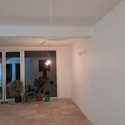 TARGET INC SPÓŁKA Z OGRANICZONĄ ODPOWIEDZIALNOŚCIĄ - Wnętrze po malowaniu na biało, widoczne gładkie ściany, duże okno z roślinami w donicach. Wiszące kable elektryczne czekają na podłączenie.