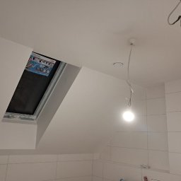 TARGET INC SPÓŁKA Z OGRANICZONĄ ODPOWIEDZIALNOŚCIĄ - Białe, świeżo pomalowane poddasze z oknem dachowym i wiszącą żarówką. Widoczne płytki na ścianie i fragment instalacji elektrycznej.