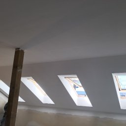 TARGET INC SPÓŁKA Z OGRANICZONĄ ODPOWIEDZIALNOŚCIĄ - Wnętrze poddasza po remoncie: gładkie, białe ściany i sufit z oknami dachowymi. Widoczny drewniany słup i fragmenty ścian w trakcie prac wykończeniowych.