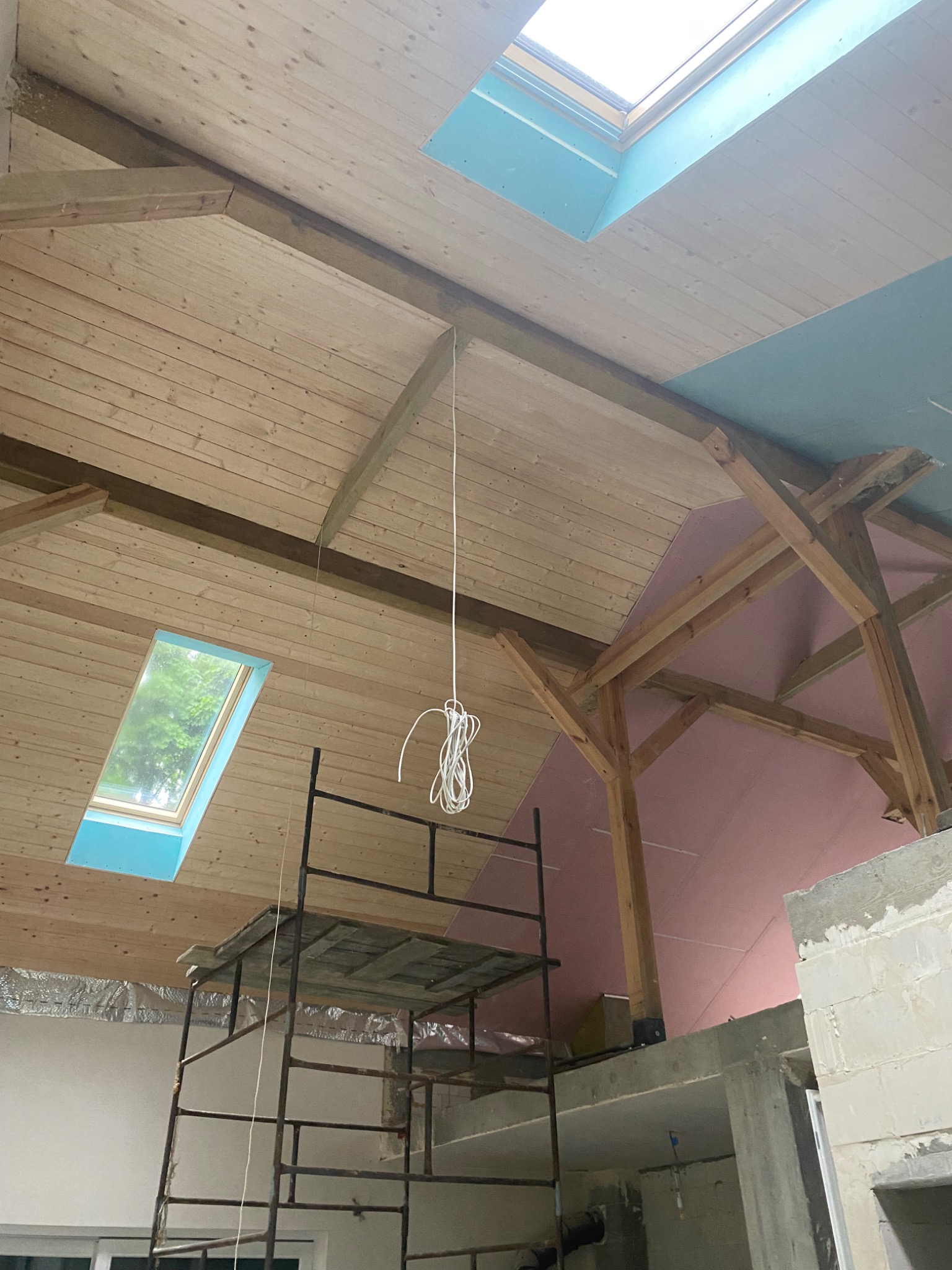 Drewniany strop z oknami dachowymi i fragmentem szarej płyty karton-gips. Widoczne rusztowanie. Ujęcie z dołu na poddaszu w trakcie prac wykończeniowych.