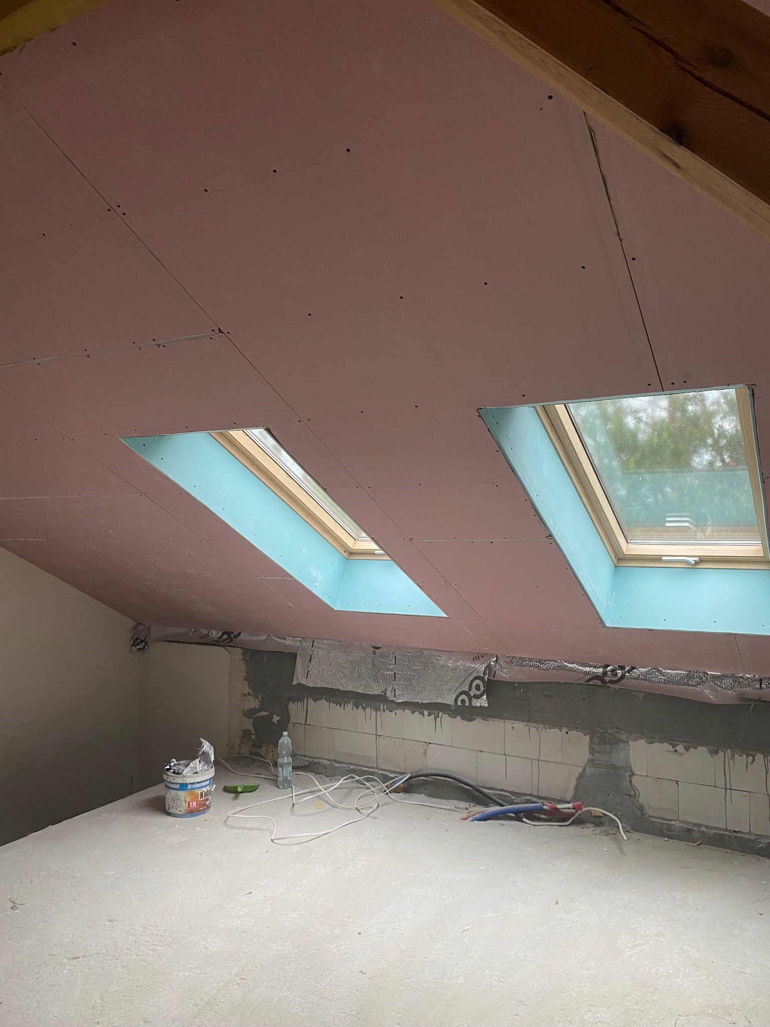 Montaż różowych płyt G-K na poddaszu ze świetlikami. Widoczne łączenia i śruby. Na podłodze resztki materiałów budowlanych i narzędzia.