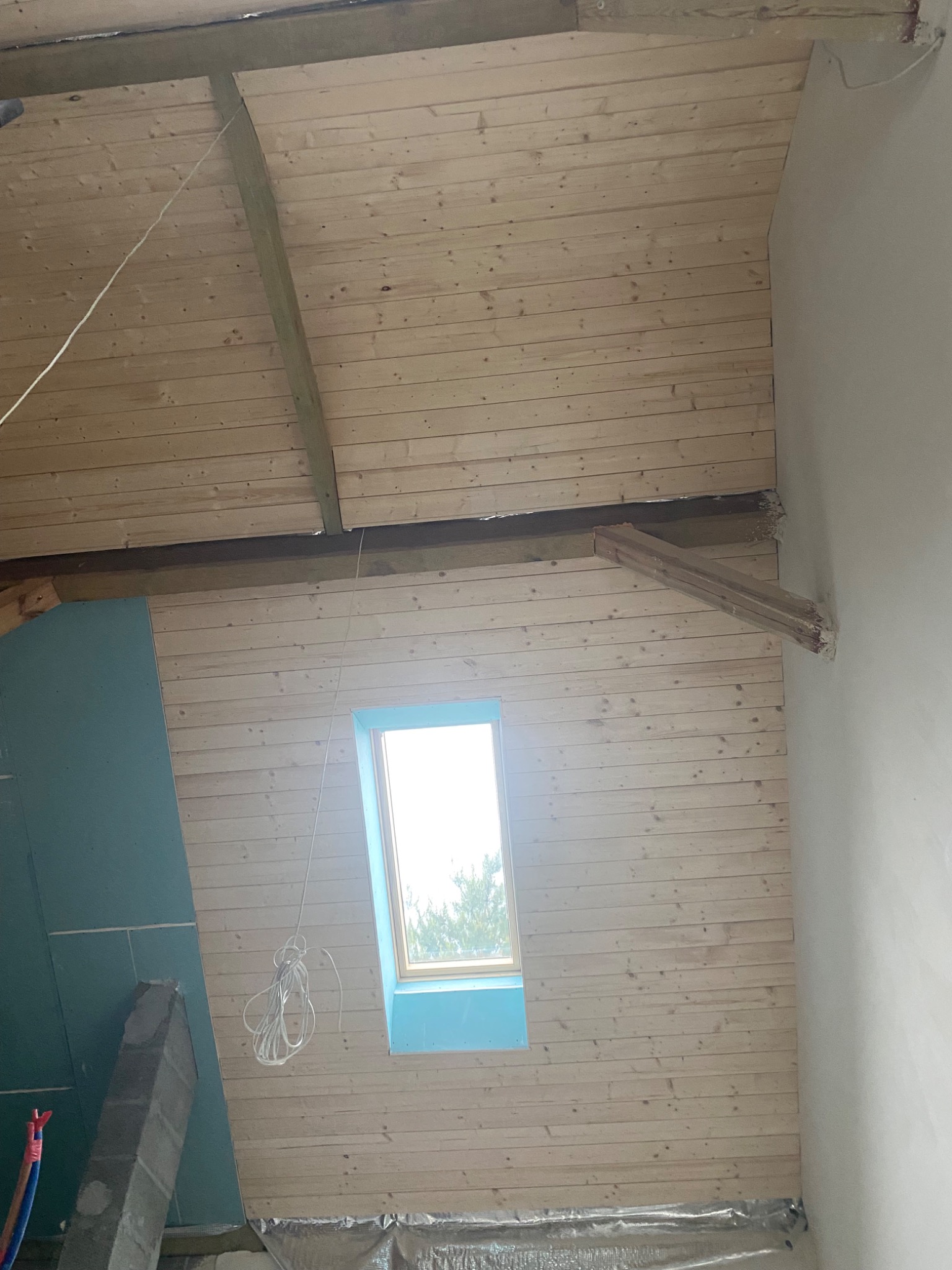 Montaż boazerii na poddaszu w Warszawie. Widoczne drewniane panele, okno dachowe i fragment ściany z płytą karton-gips. Prace wykończeniowe w trakcie realizacji.