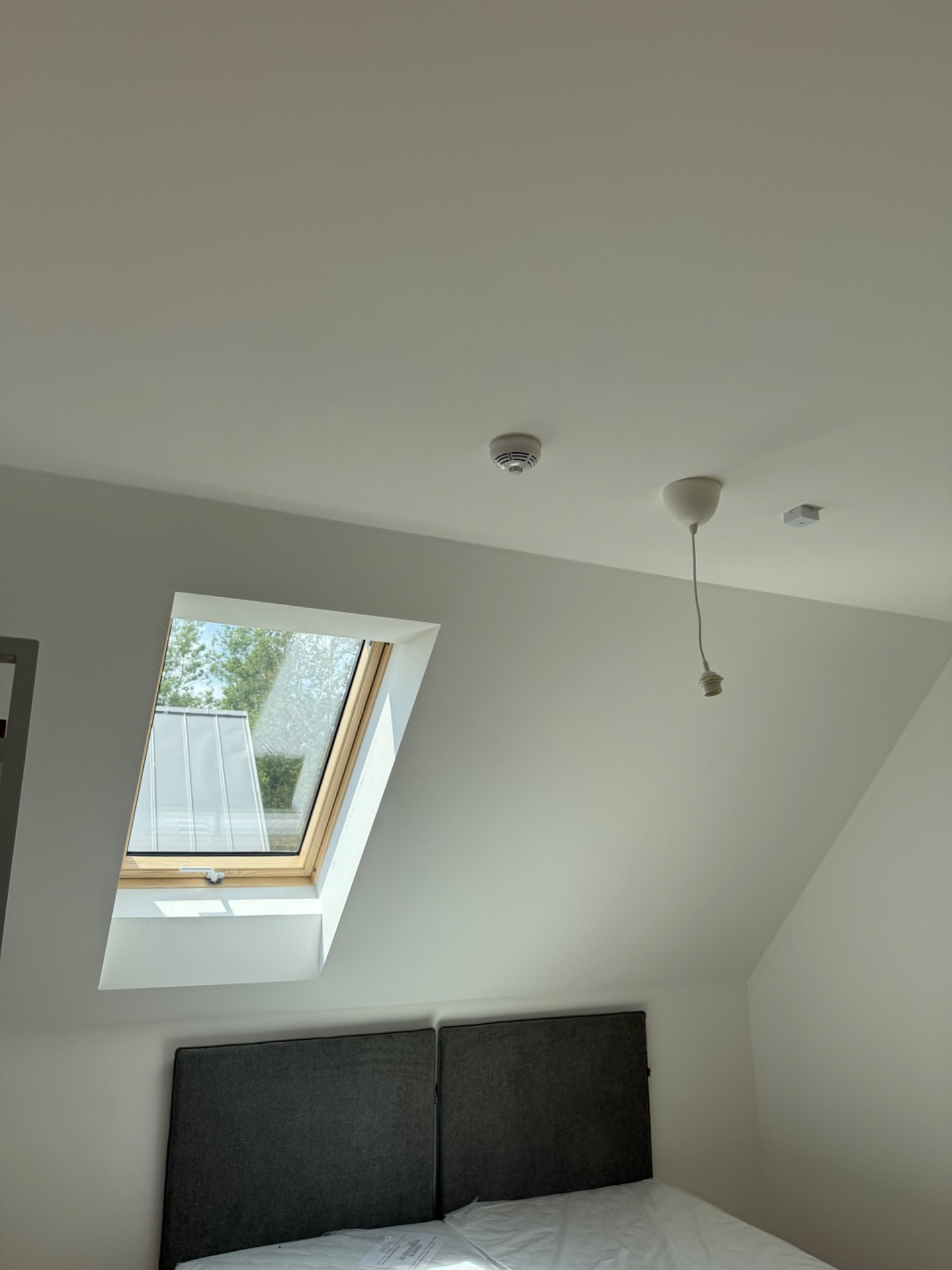 Poddasze po remoncie: białe ściany i sufit, okno dachowe z widokiem na zewnątrz, czujnik dymu, oprawa oświetleniowa bez żarówki i szare wezgłowie łóżka.