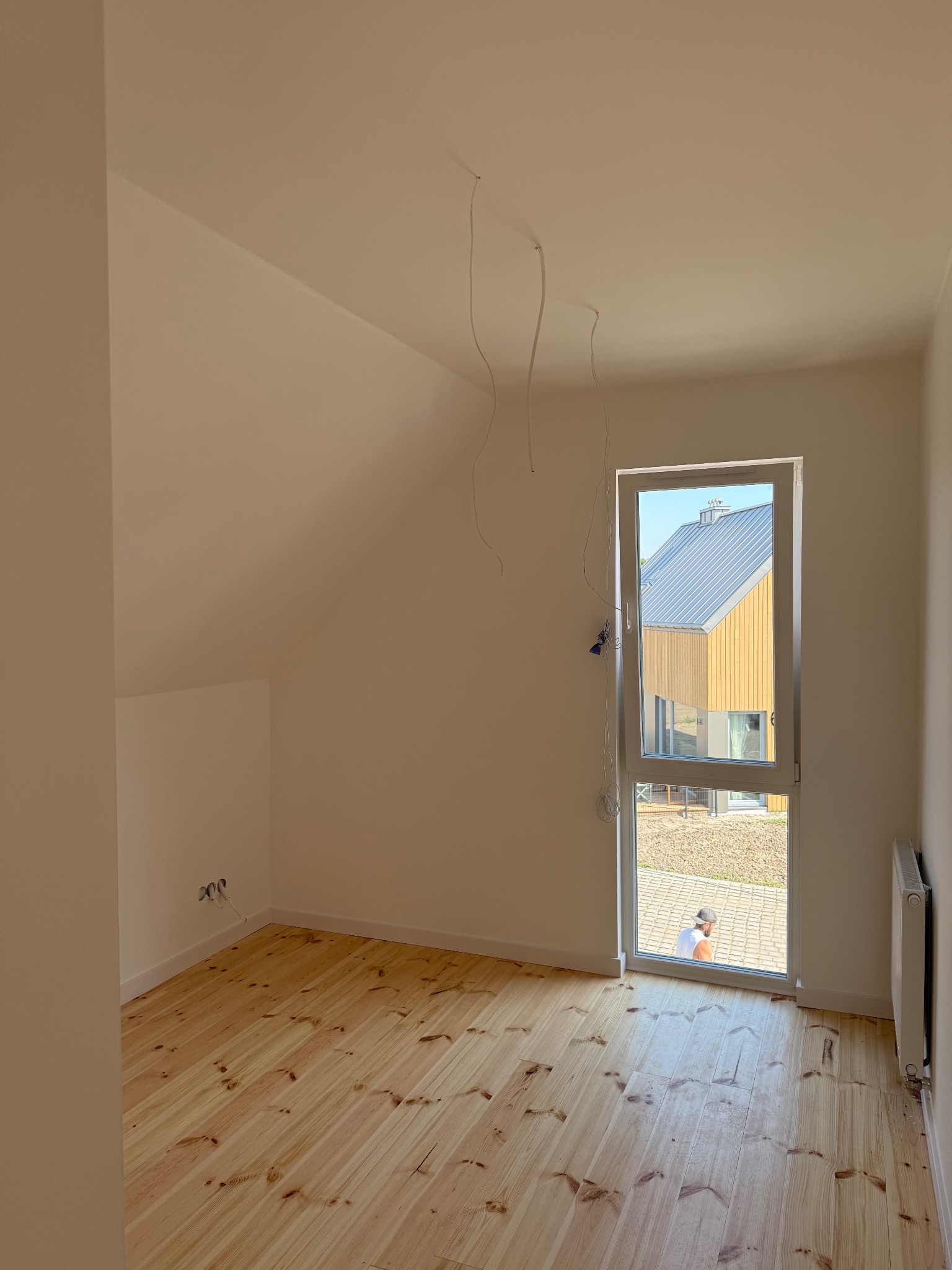 Jasne wnętrze poddasza z drewnianą podłogą i oknem, przez które widać nowoczesny dom. Ściany pomalowane na biało, widoczne kable wystające z sufitu.