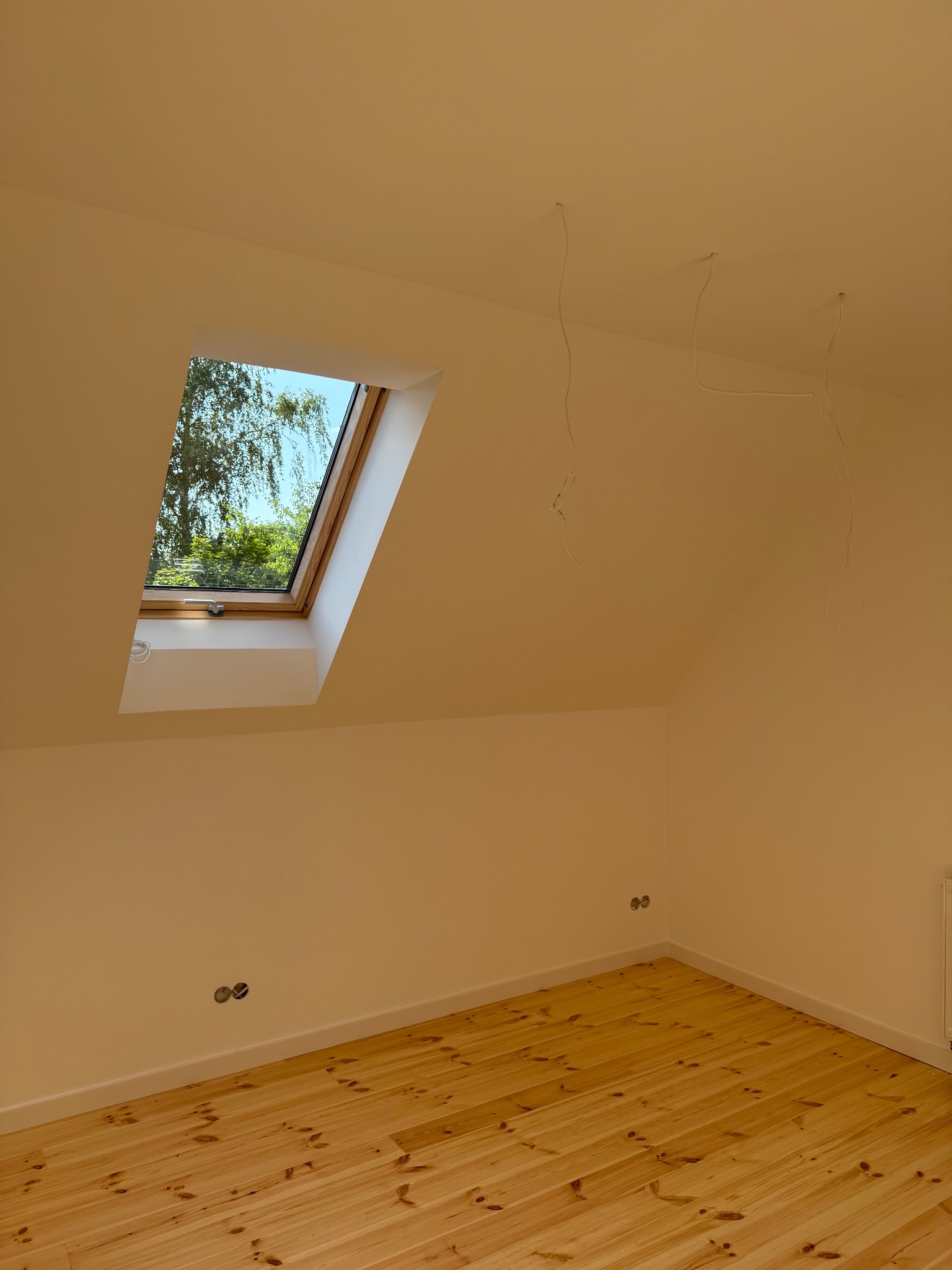 Jasne wnętrze po malowaniu z drewnianą podłogą i oknem dachowym, przez które widać drzewa. Z sufitu zwisają kable.