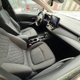 MarClean Pranie Tapicerek - Wnętrze samochodu z szarą tapicerką foteli, widoczne detale deski rozdzielczej, kierownicy i ekranu dotykowego, szyba przednia pokryta kroplami deszczu.