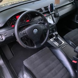 MarClean Pranie Tapicerek - Wnętrze samochodu Volkswagen Passat z czarną tapicerką, widoczny kokpit, kierownica z logo VW, skórzane fotele z elementami z alcantary i wisząca zapachowa zawieszka 'Fresso'.