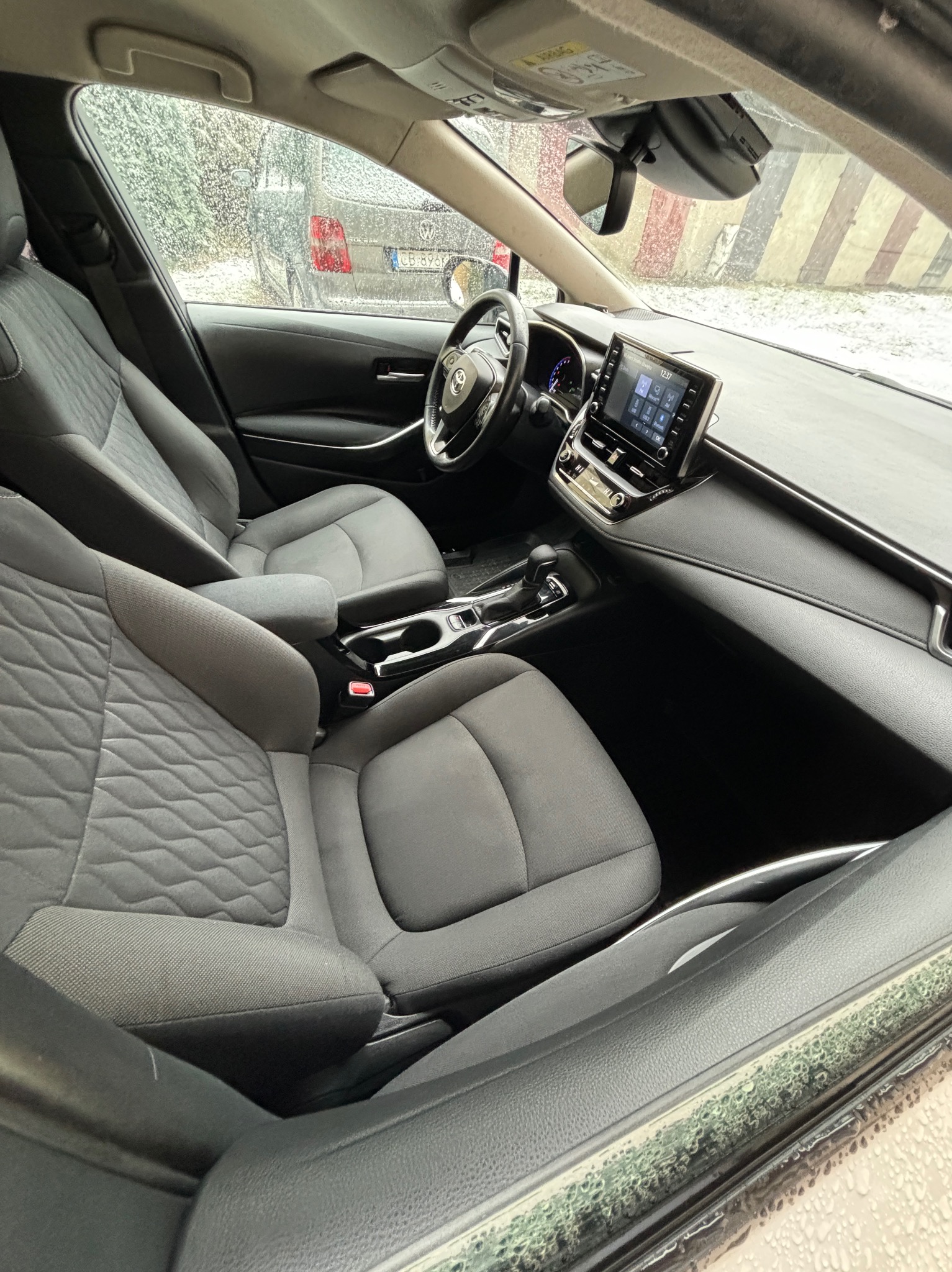 Wnętrze samochodu z szarą tapicerką foteli, widoczne detale deski rozdzielczej, kierownicy i ekranu dotykowego, szyba przednia pokryta kroplami deszczu.