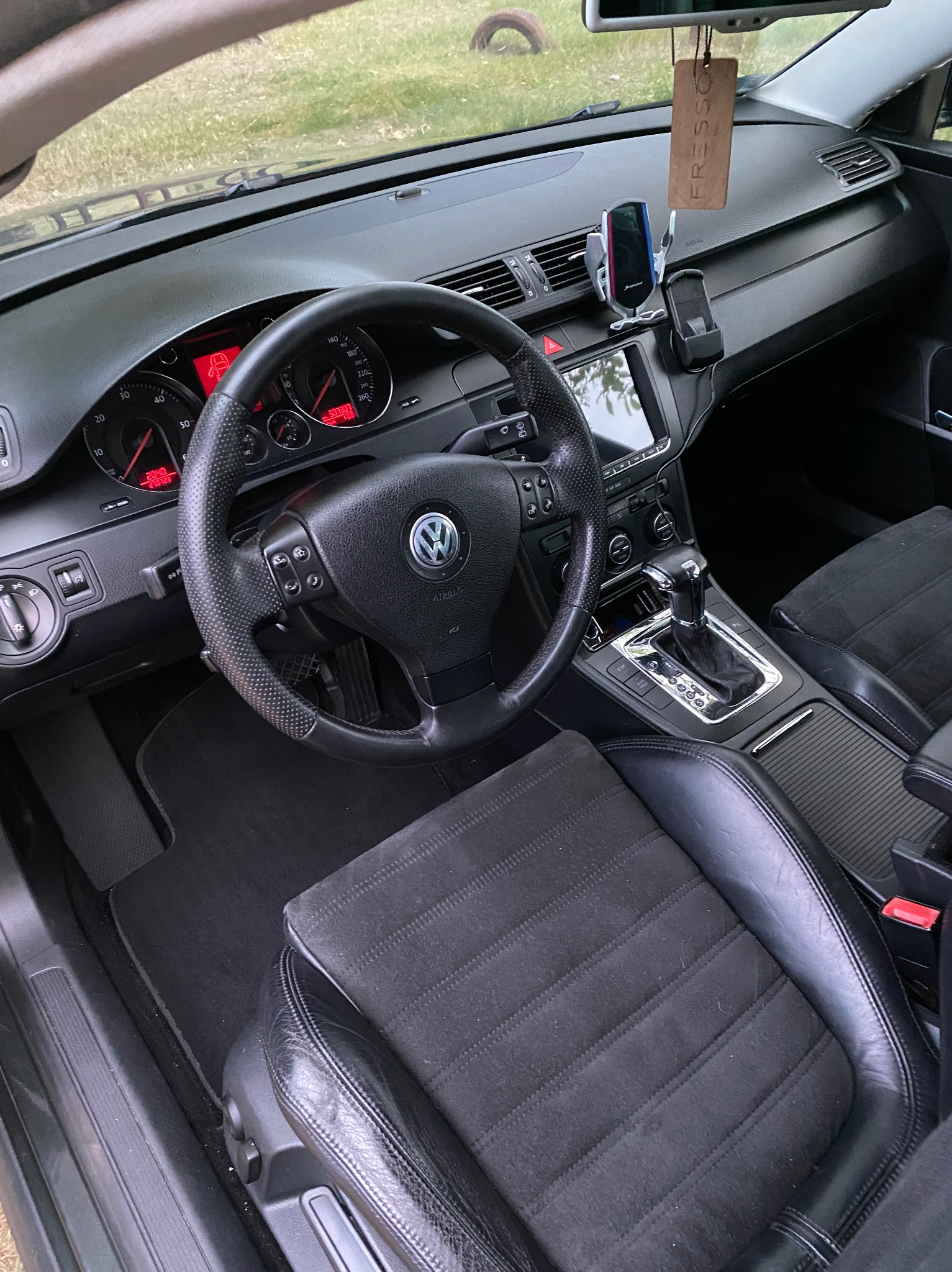 Wnętrze samochodu Volkswagen Passat z czarną tapicerką, widoczny kokpit, kierownica z logo VW, skórzane fotele z elementami z alcantary i wisząca zapachowa zawieszka 'Fresso'.